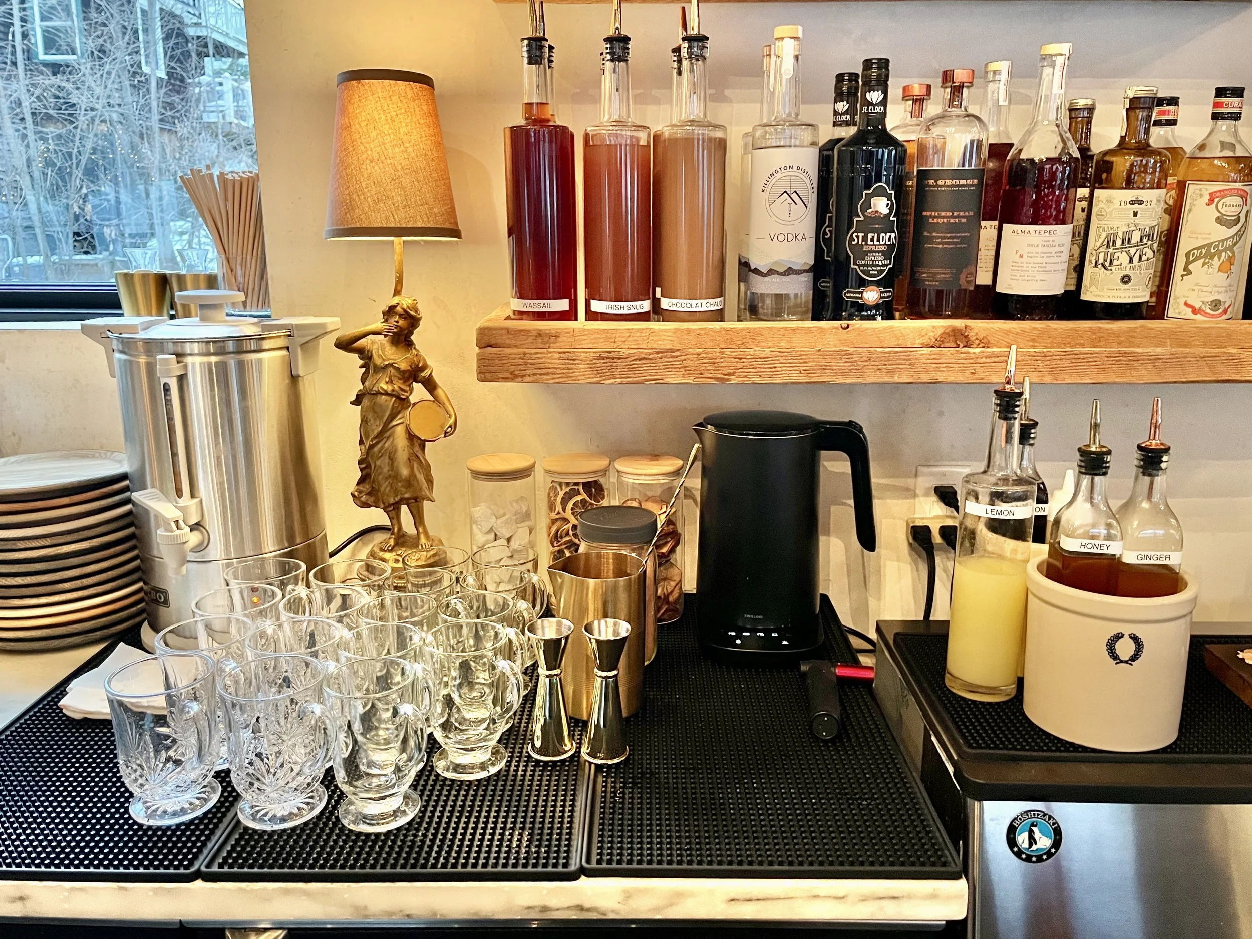 Larger mugs left, smaller mugs right. 3 garnish jars along back: marshmallow, lemon, apple. Kettle is pushed all the way back near garnishes. Cocoa powder jar with tbsp inside, mixing glass, jiggers, and measuring cup (not pictured) lined up beside g