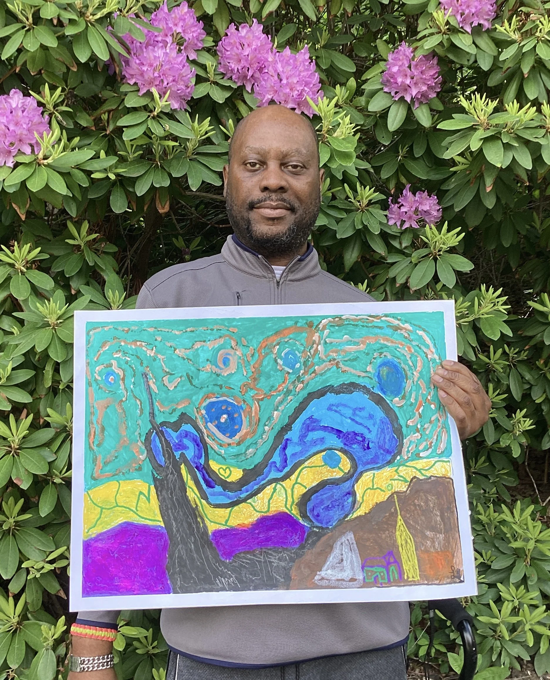 A man standing in front of greenery with pink flowers, holding a colorful abstract painting.