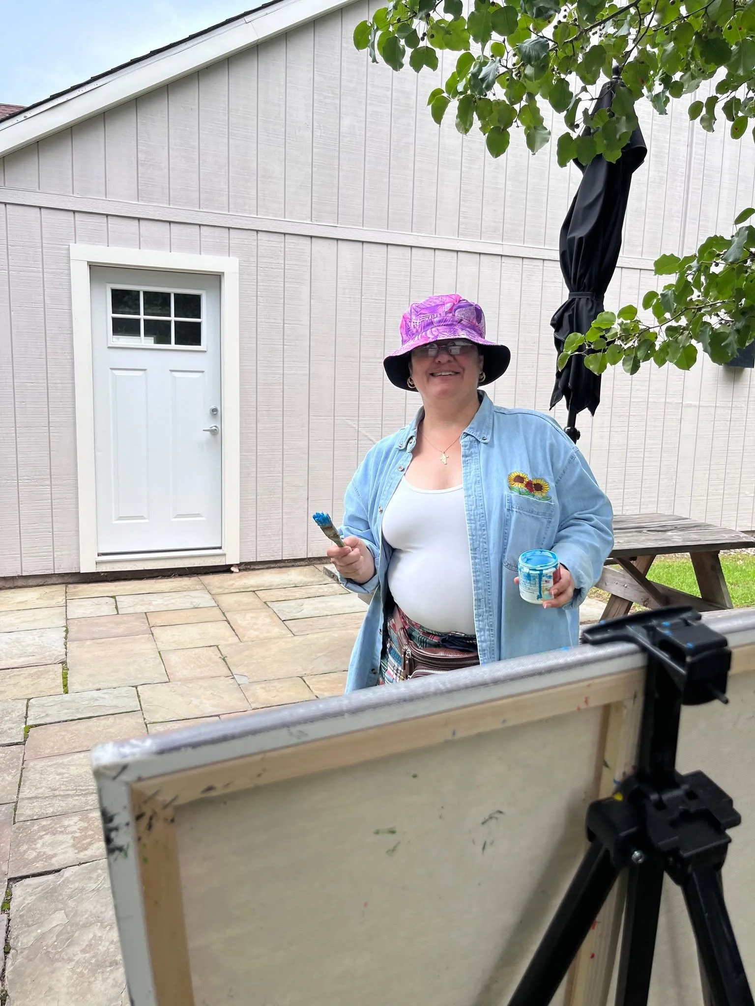 A woman wearing a purple hat and glasses holds a paintbrush and a small container, standing outside in front of a beige house with a white door and an outdoor umbrella. She is smiling and wearing a light blue shirt with a sunflower patch.