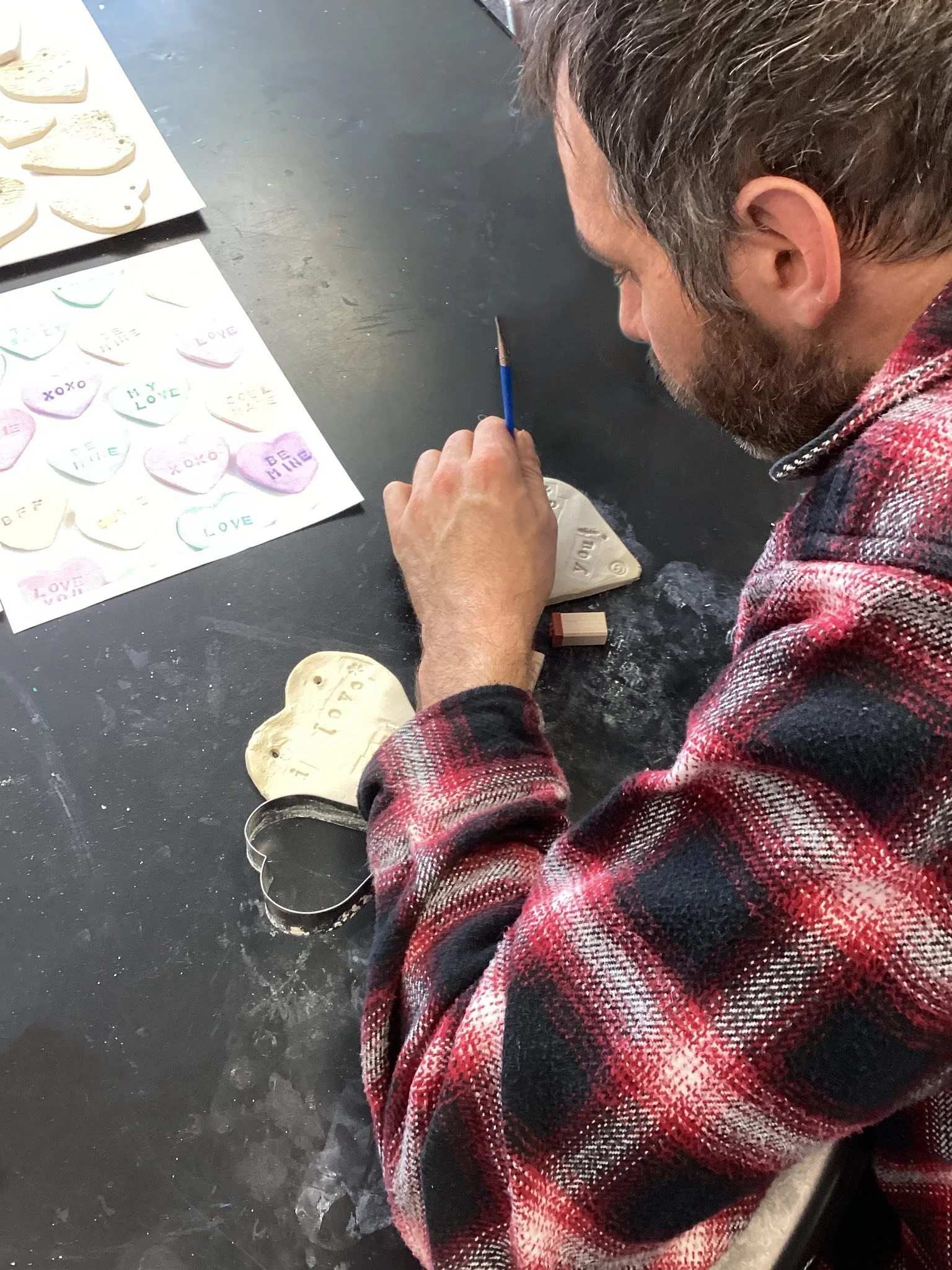 A man with gray hair and beard wearing a red and black plaid shirt sitting at a black table. He's holding a wooden stamp in his right hand and pressing it onto a circular wooden ornament that has the phrase "LOVE" carved into it. On the table, to his left, there is a sheet with various heart-shaped paper cutouts featuring words like "LOVE," "XOXO," and "BE MINE." There is also a tray with several heart-shaped cookies or crafts.