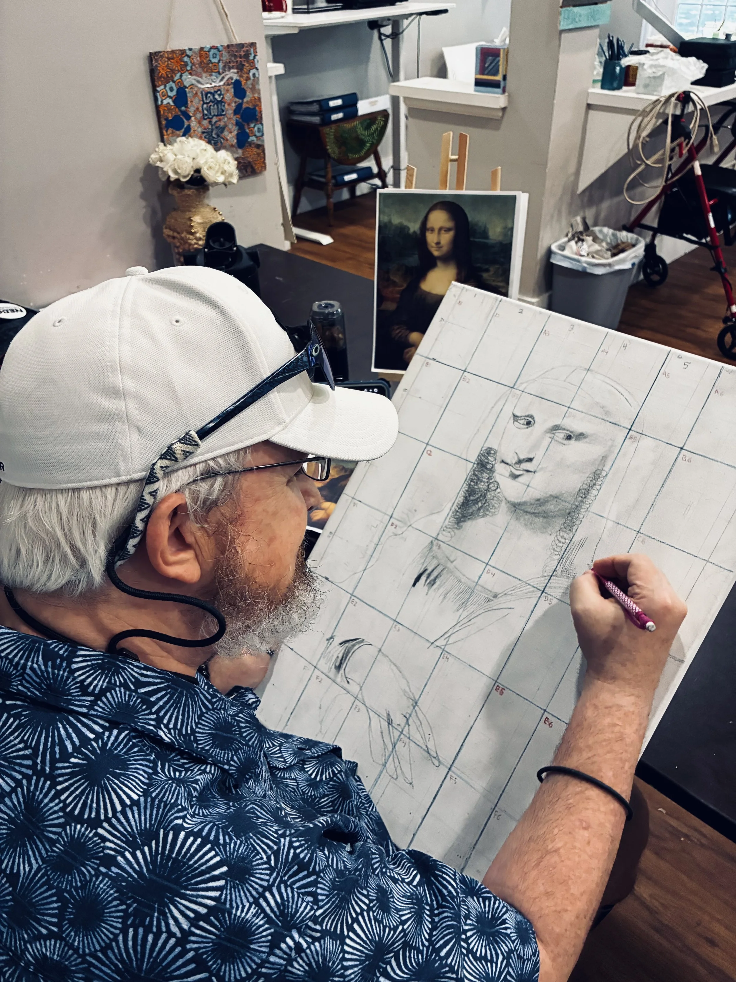 An elderly man in a patterned blue shirt and white cap drawing a portrait of the Mona Lisa on graph paper. He is holding a pink pen, and his glasses are on his nose. Behind the man, there is a table with various art supplies and a printed image of the Mona Lisa on an easel.