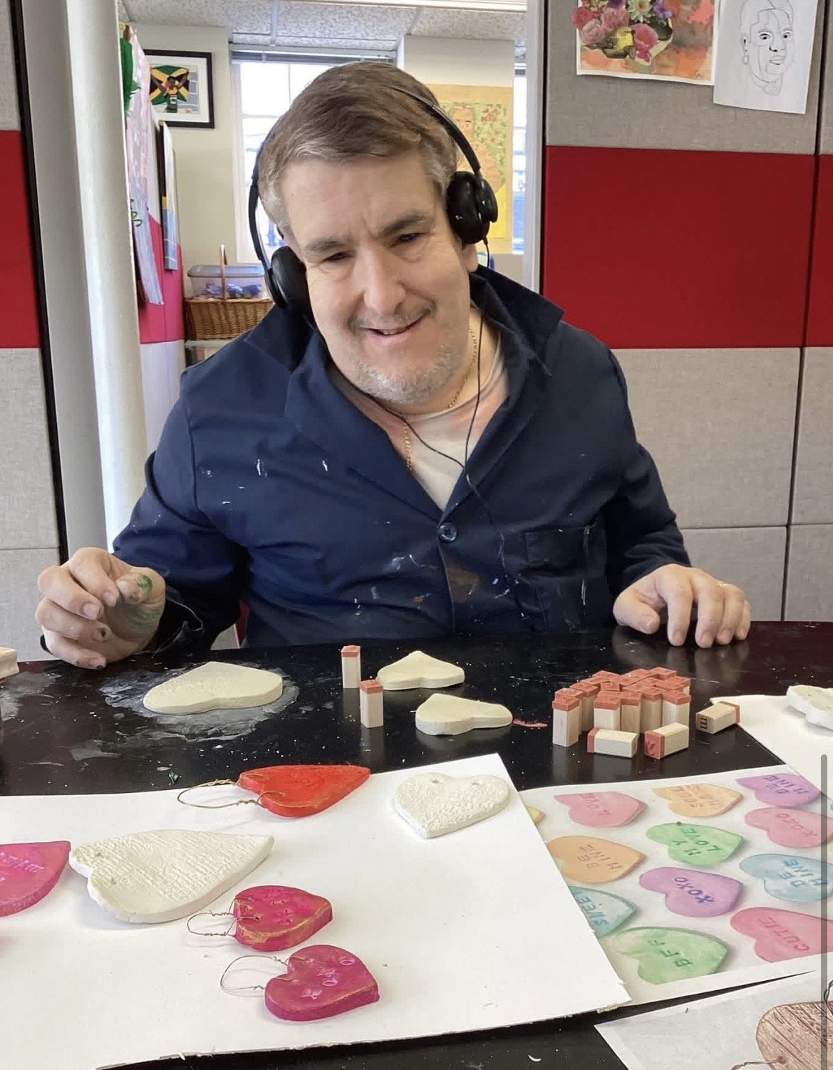 A man with a headset, smiling, sitting at a table with various heart-shaped and other shaped sugar cookies decorated with colored icing and words, along with small wooden blocks and a sheet with colorful heart shapes and messages.