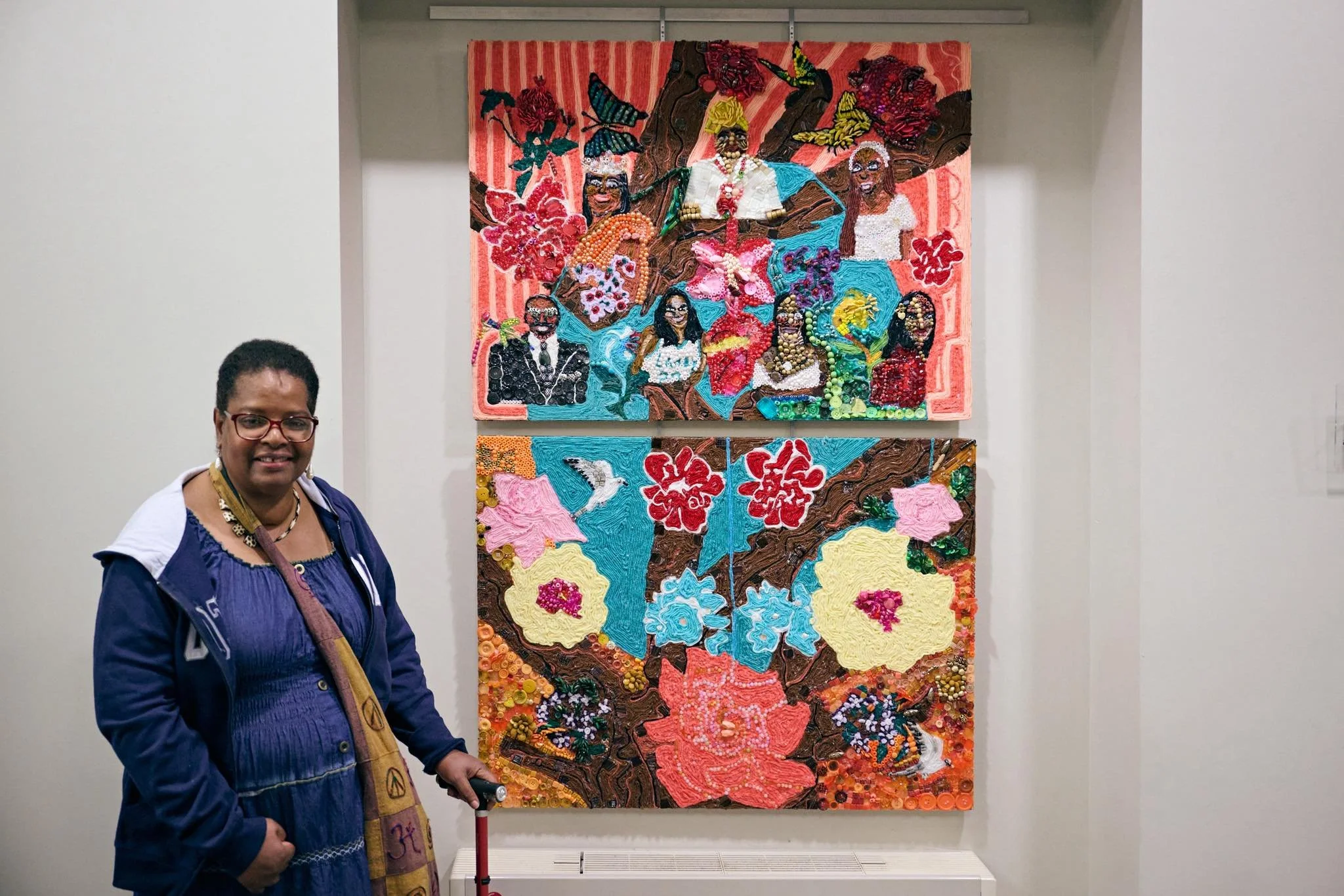 A woman with glasses, earrings, and a blue jacket stands in front of a colorful quilted artwork depicting African American women with jewelry, headwraps, and natural hair, holding hands with a background of butterflies, flowers, and leaves.