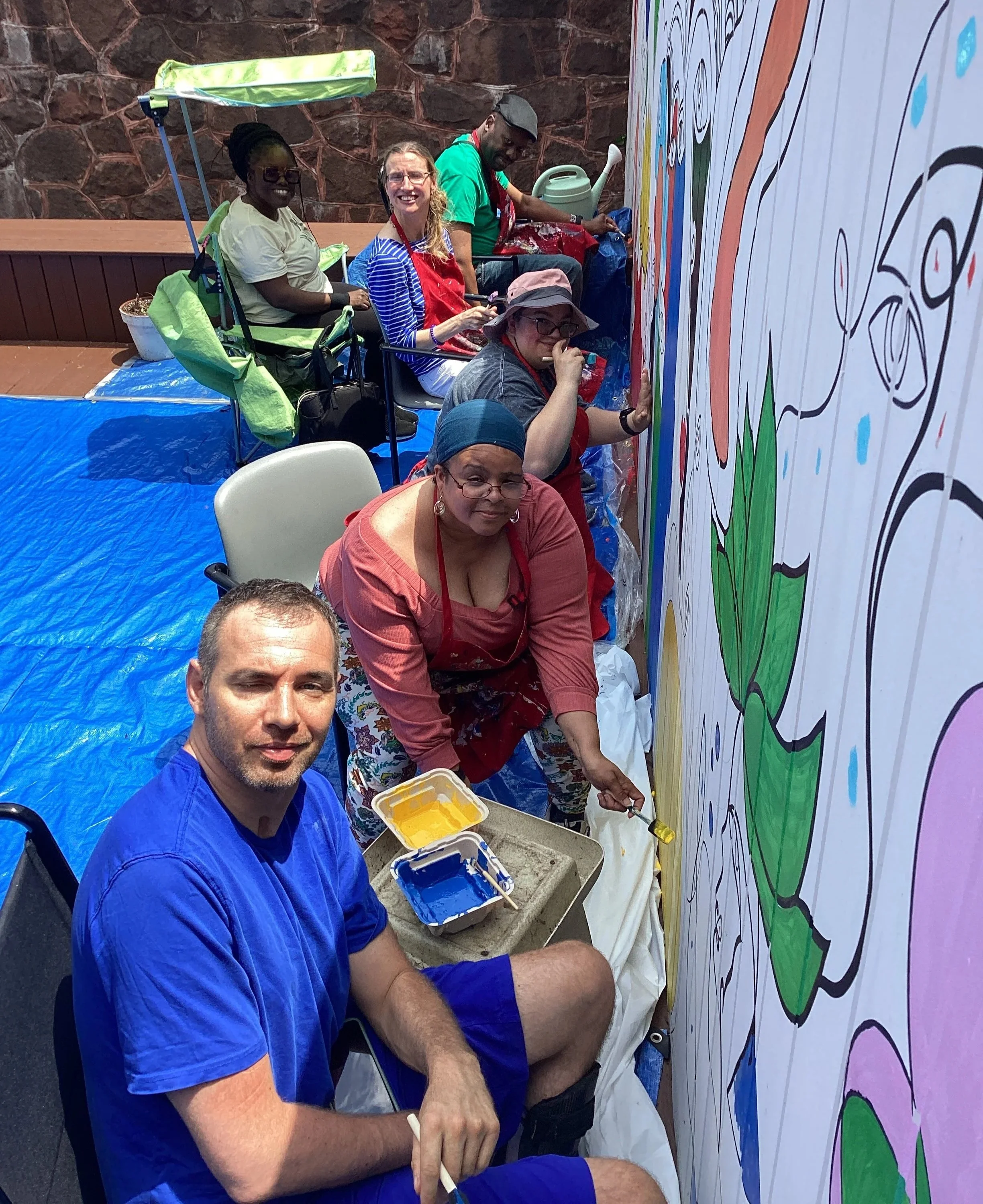 Group of people painting a large colorful mural on a wall outdoors, some sitting, some standing, with painting supplies nearby.