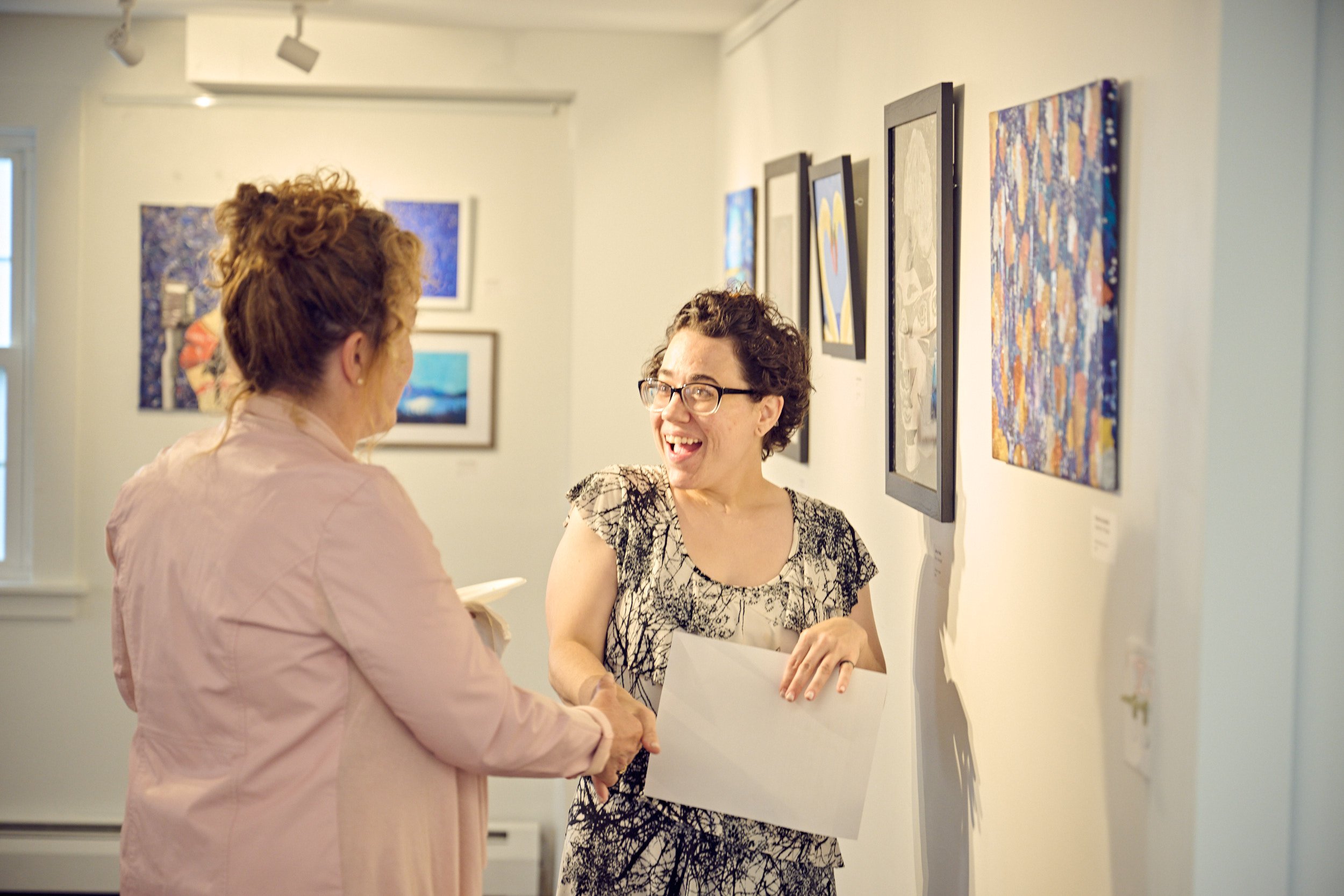 Two women smiling and shaking hands at an art gallery, with colorful paintings hanging on the wall behind them.