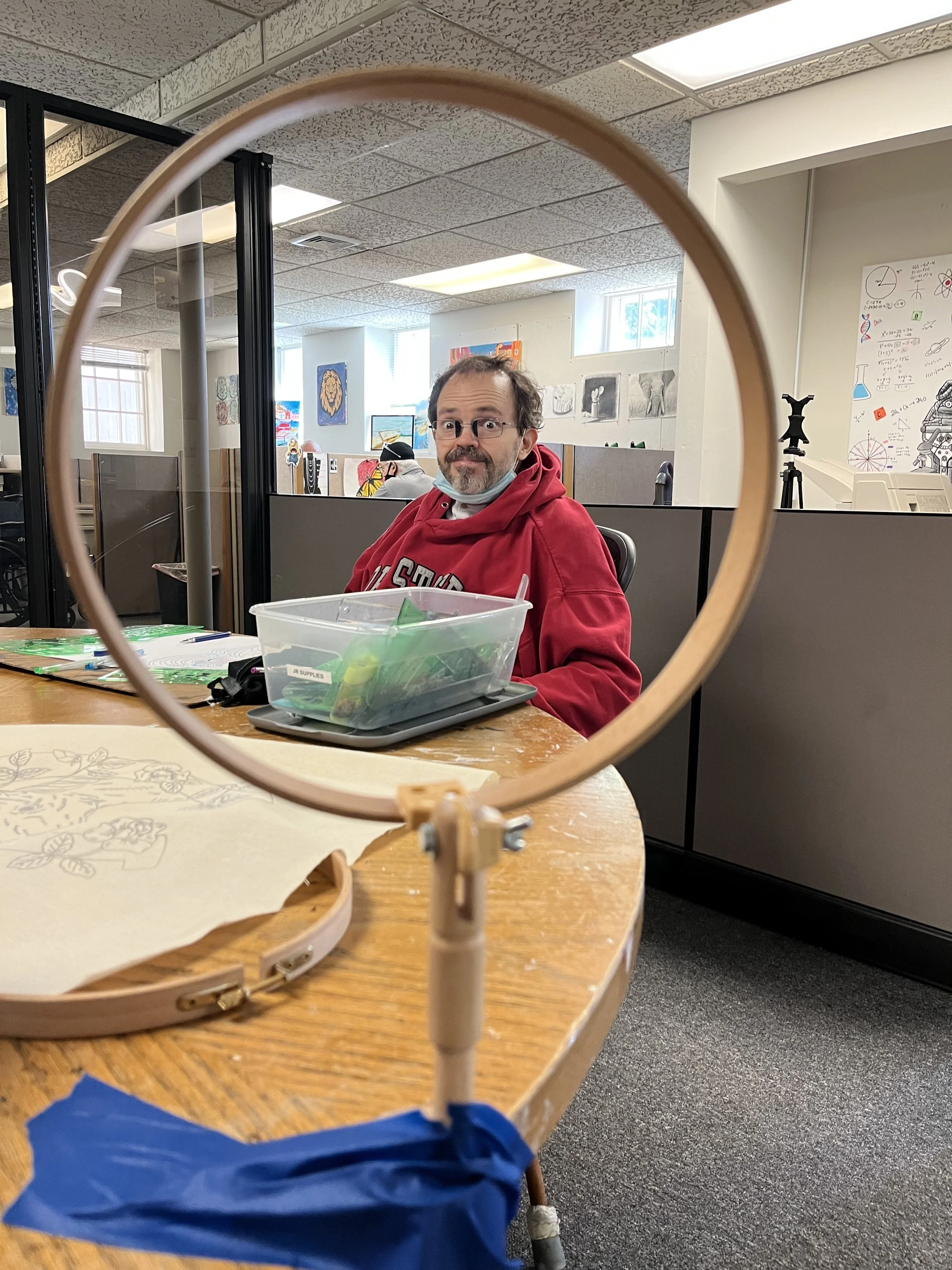 A man in a red hoodie and glasses sitting at a table in an office, seen through a wooden embroidery hoop on the table.