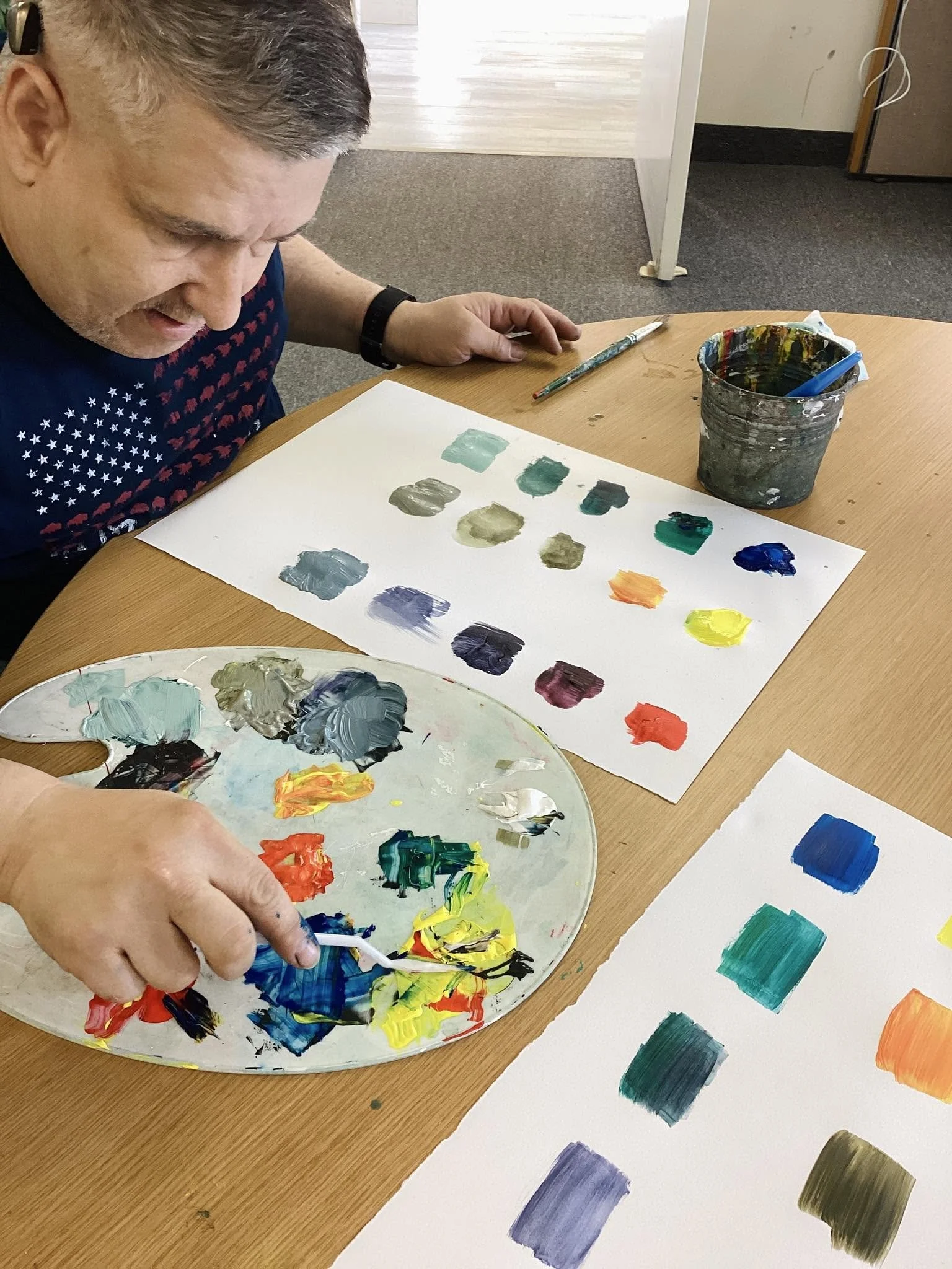 Person mixing different paint colors on a palette while working on a color chart or painting at a wooden table.