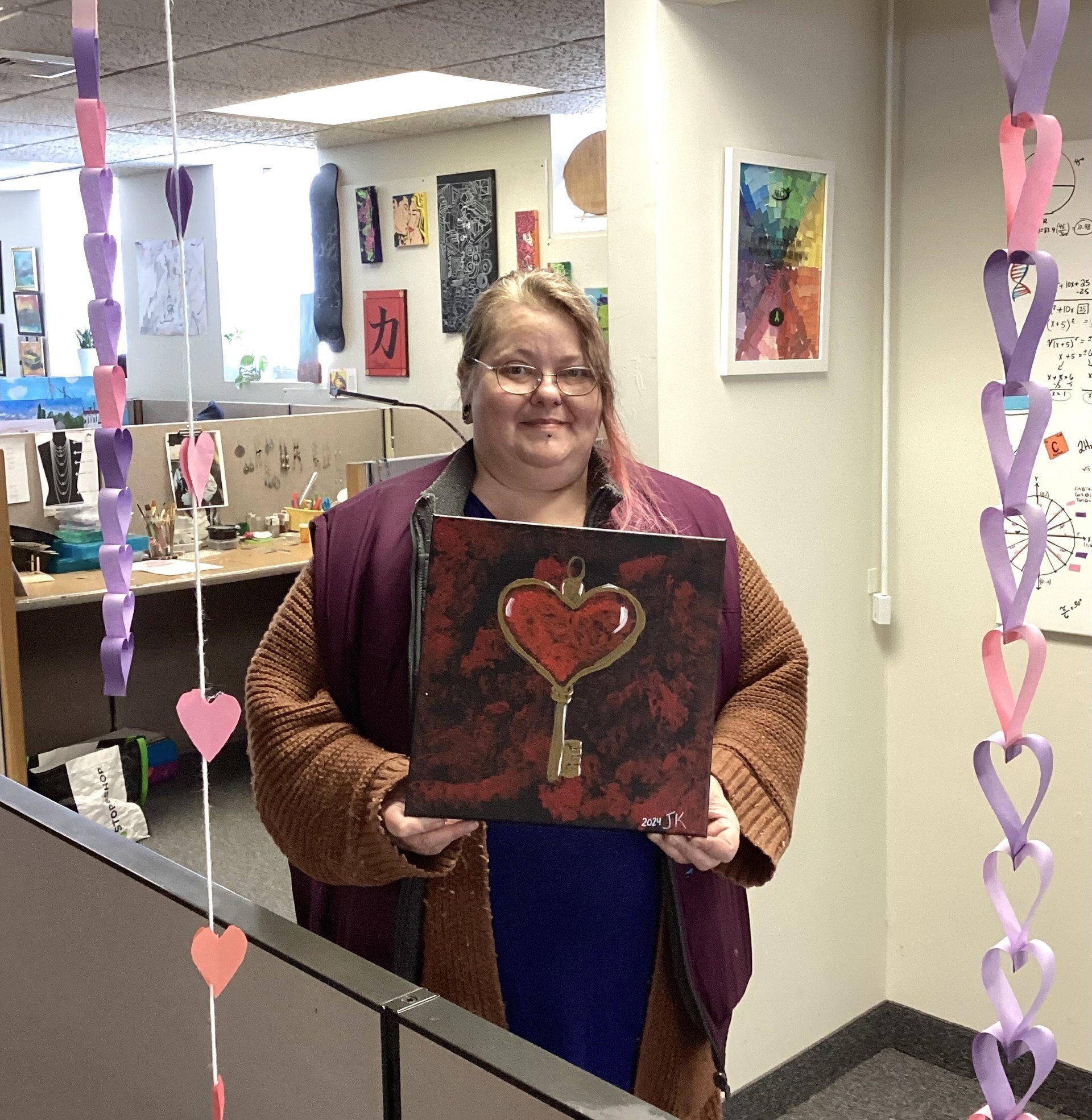 A woman with glasses and light brown hair, wearing a burgundy jacket over a brown sweater, stands inside an office holding a painting of a red heart outlined in gold. There are multicolored paper hearts hanging on ribbons in front of her, and the office has cubicles, artwork on the walls, and a whiteboard with mathematical equations.
