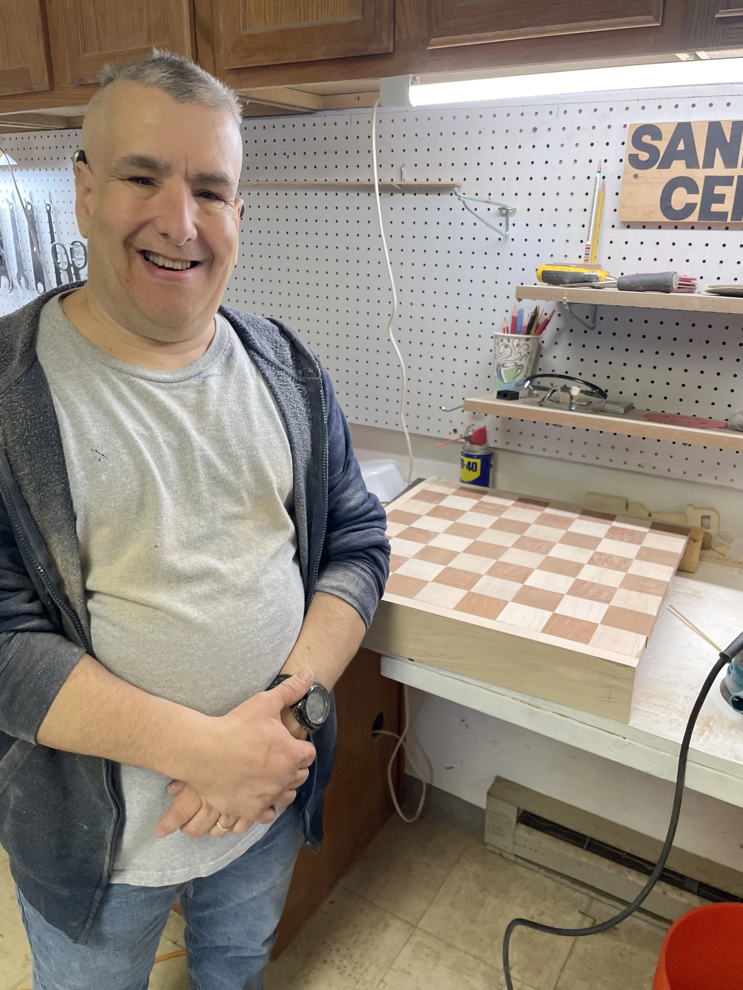 Smiling man standing next to a woodworking project in a workshop with pegboard wall, tools, and shelves, holding his wrist.