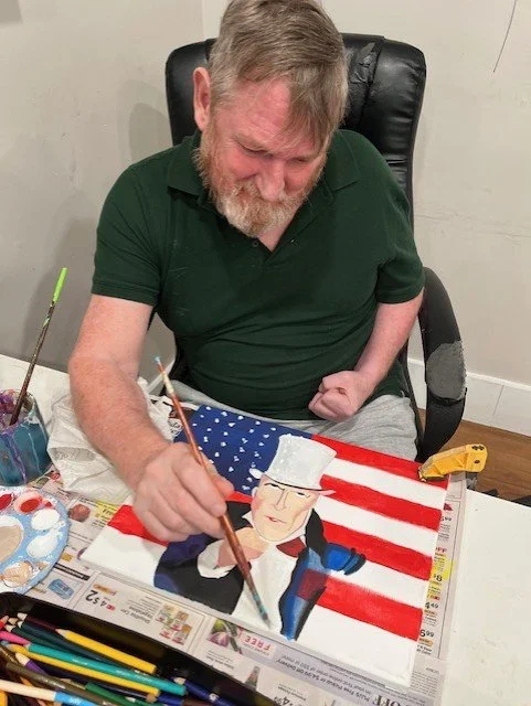 An elderly man with gray hair and a beard, wearing a dark green polo shirt, is sitting at a table painting a portrait of Donald Trump on a canvas. The background features an American flag design, and the man appears focused and happy.