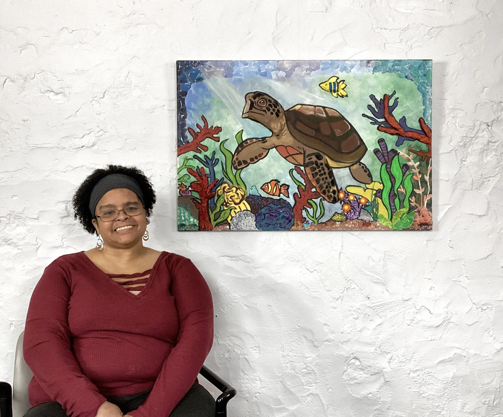 Woman sitting in a chair in front of a colorful underwater-themed painting on a white brick wall.