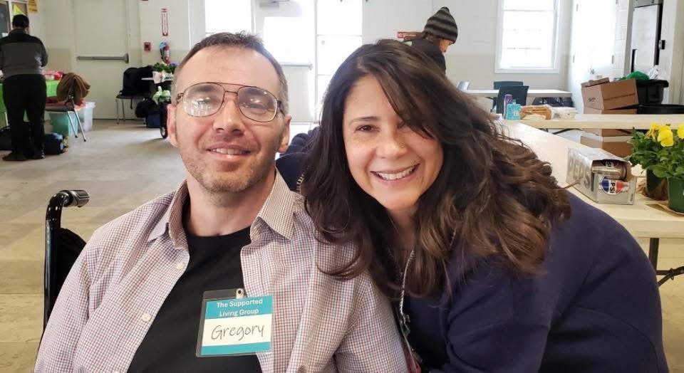 A man and a woman smiling at the camera, sitting close together indoors. The man is wearing glasses and a name tag that says 'Gregory'. The woman has long dark hair and is wearing a dark jacket. In the background, there are tables, boxes, and a person wearing a beanie.