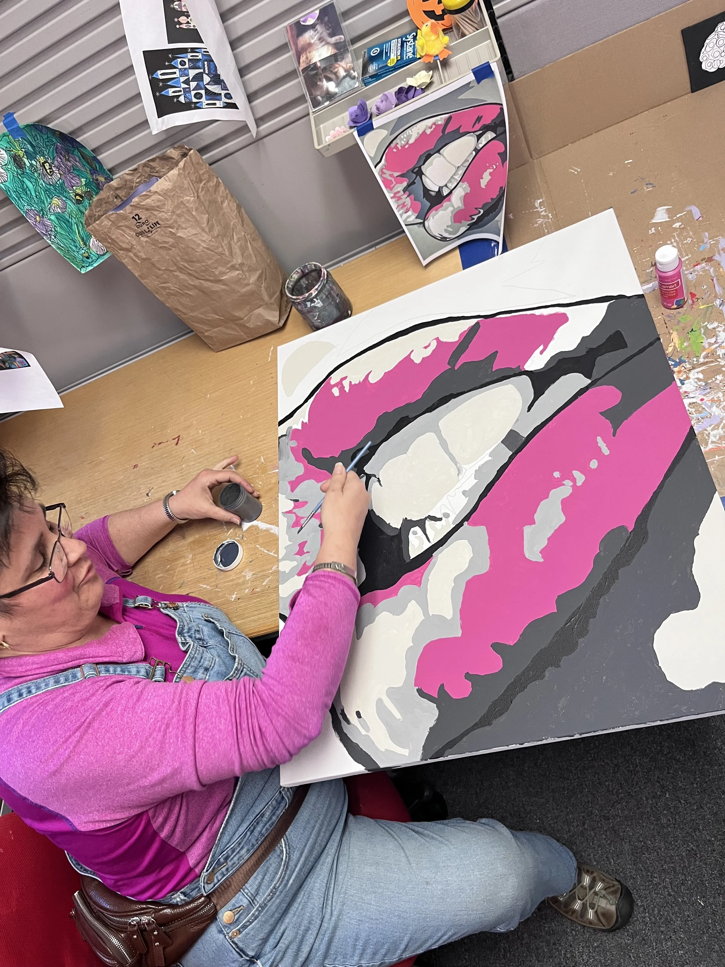 A woman wearing a pink top, denim vest, and jeans is painting a canvas featuring a stylized, close-up image of lips with pink lipstick and teeth. The workspace has art supplies, including paint jars and printed references of similar artwork.