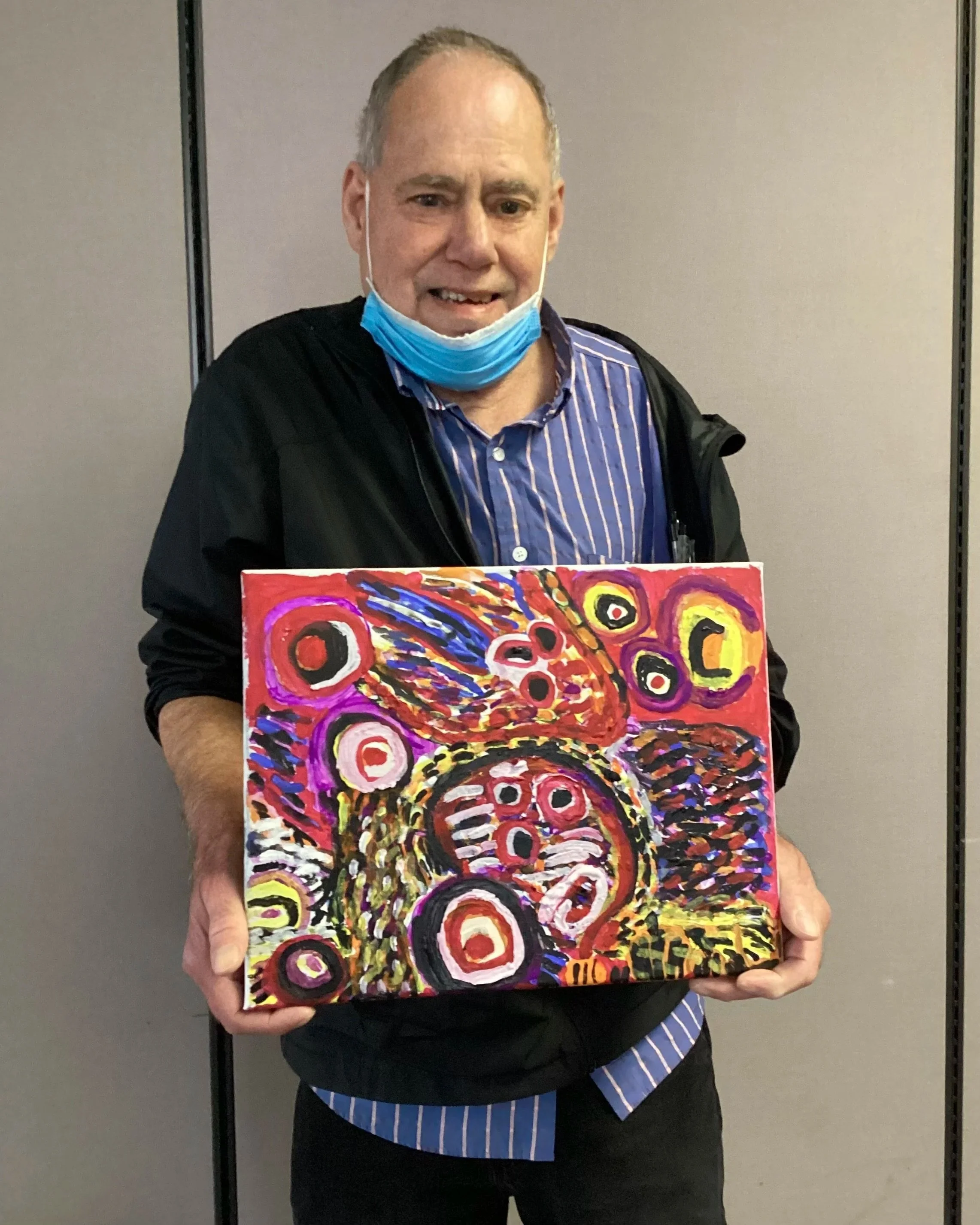 A man wearing a blue striped shirt, black jacket, and a face mask pulled down is holding a colorful abstract painting with circular patterns in red, yellow, purple, and black, standing in front of a plain wall.