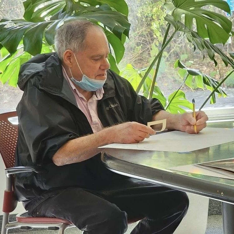An elderly man sitting at a table near a window, with large green indoor plants behind him, wearing a face mask pulled down to his chin, sketching on paper with a pencil.