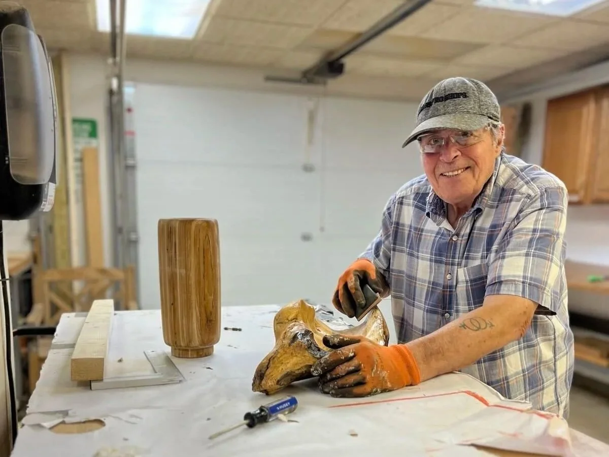 An elderly man in a plaid shirt, safety glasses, and orange gloves working on a woodworking project in a workshop. He is holding a piece of wood and smiling at the camera, with woodworking tools and materials on the table.