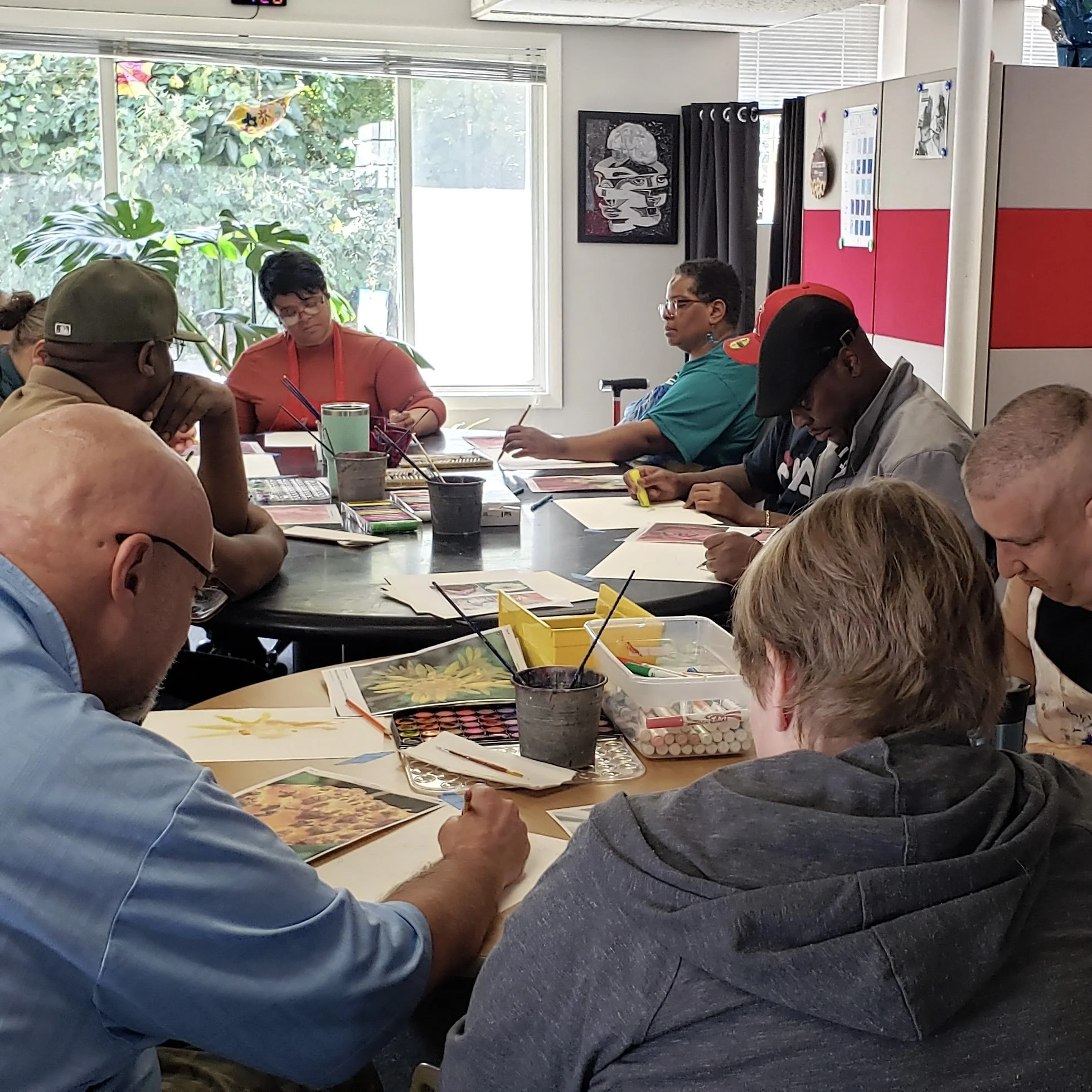 People seated around a table engaging in arts and crafts activities, with art supplies like paint palettes and brushes visible, and a window showing greenery outside.