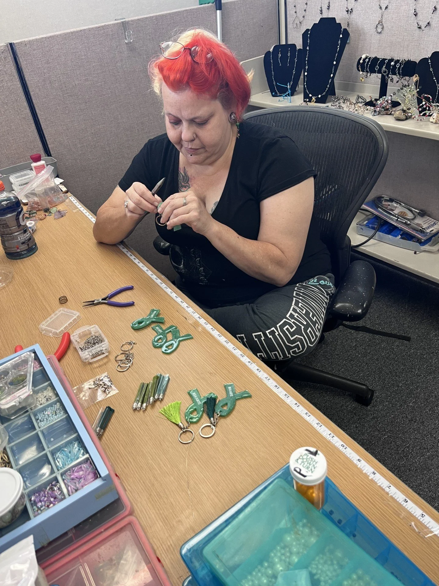 A woman with red hair and glasses on her head is crafting jewelry at a table. She has glasses on her forehead, tattoos, and is wearing a black T-shirt. The table has various jewelry-making supplies, including beads, charms, pliers, and decorative items. Behind her is a display of jewelry, with necklaces and bracelets on stands.