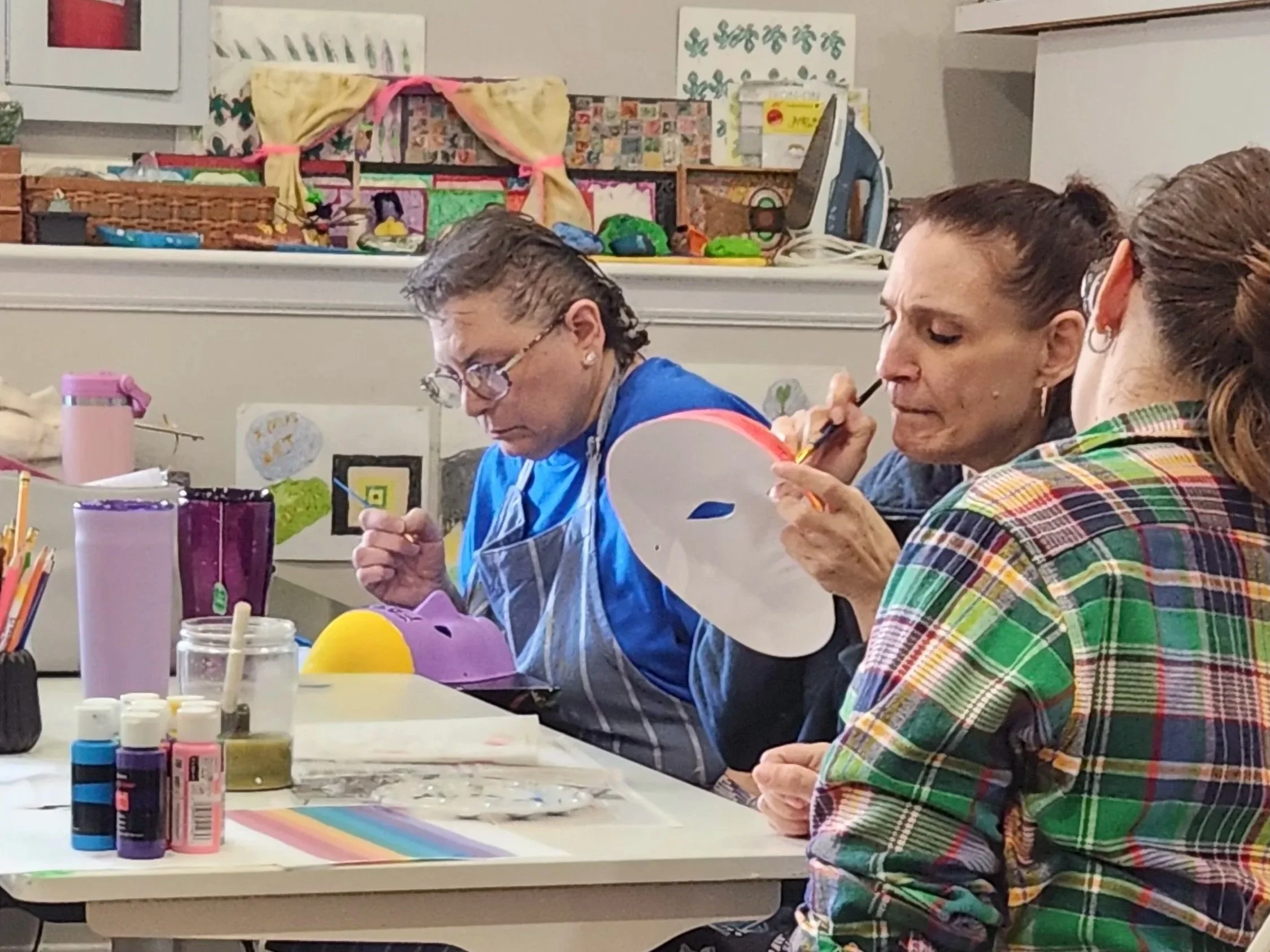 Three women sit at a long table covered with various paint supplies, including bottles of paint, brushes, and paper. They are engaged in an arts and crafts activity in a room decorated with artwork and craft displays.