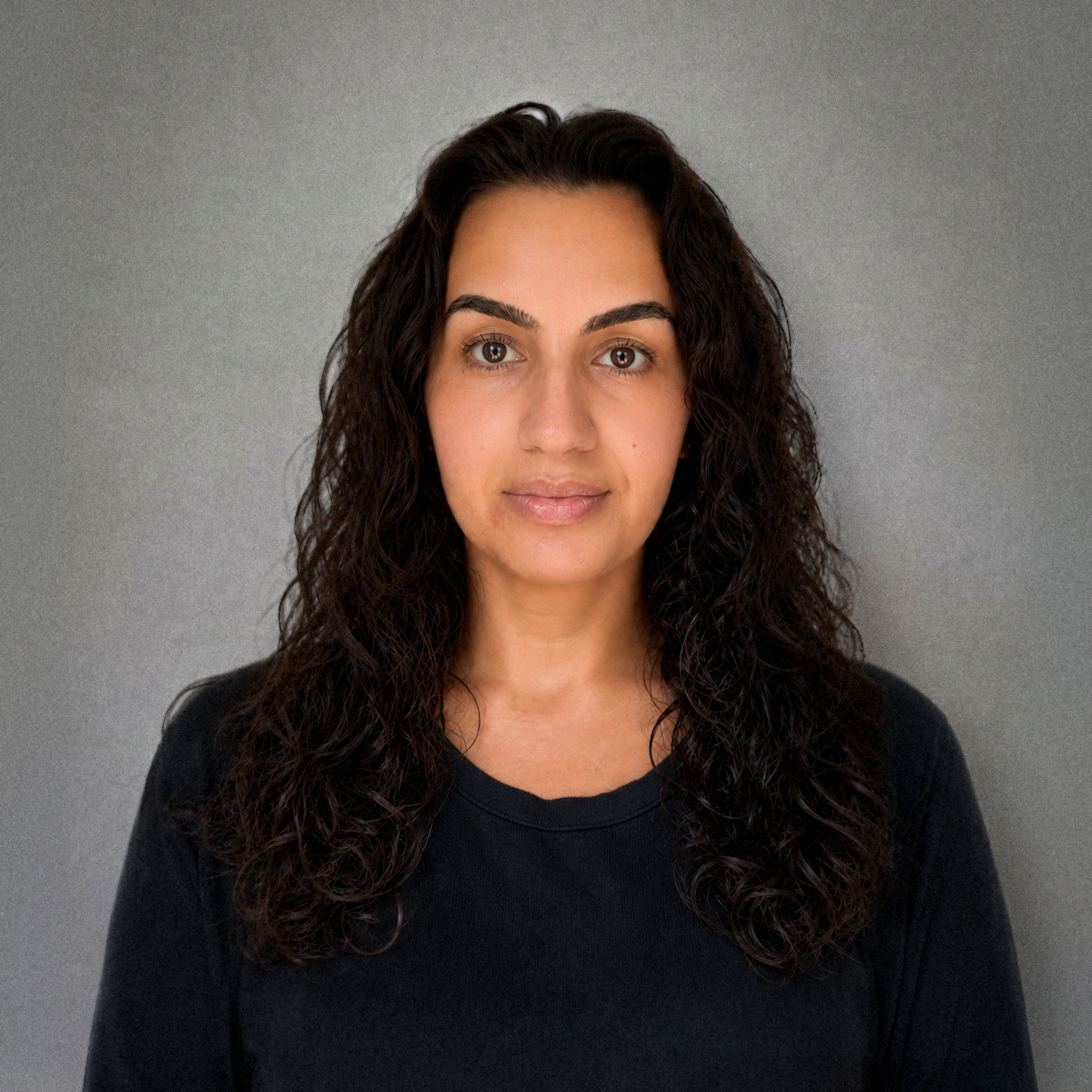 Portrait of a woman with long curly dark hair, wearing a black shirt, against a plain gray background.