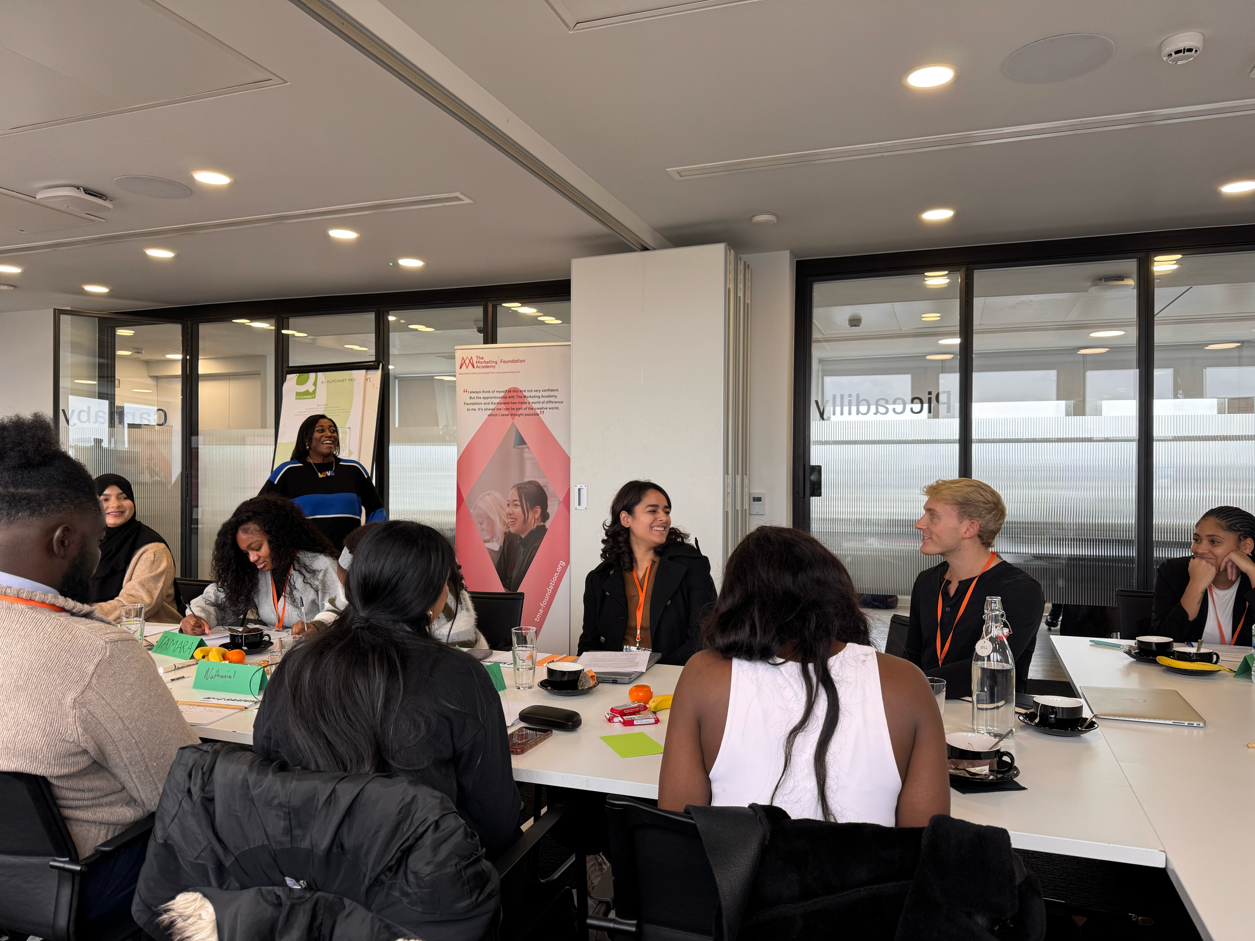 A diverse group of people gathered around a conference table in a modern office space, engaged in a discussion or presentation with a woman standing and speaking. The table has name tags, notebooks, and beverages, and there are banners and glass walls in the background.