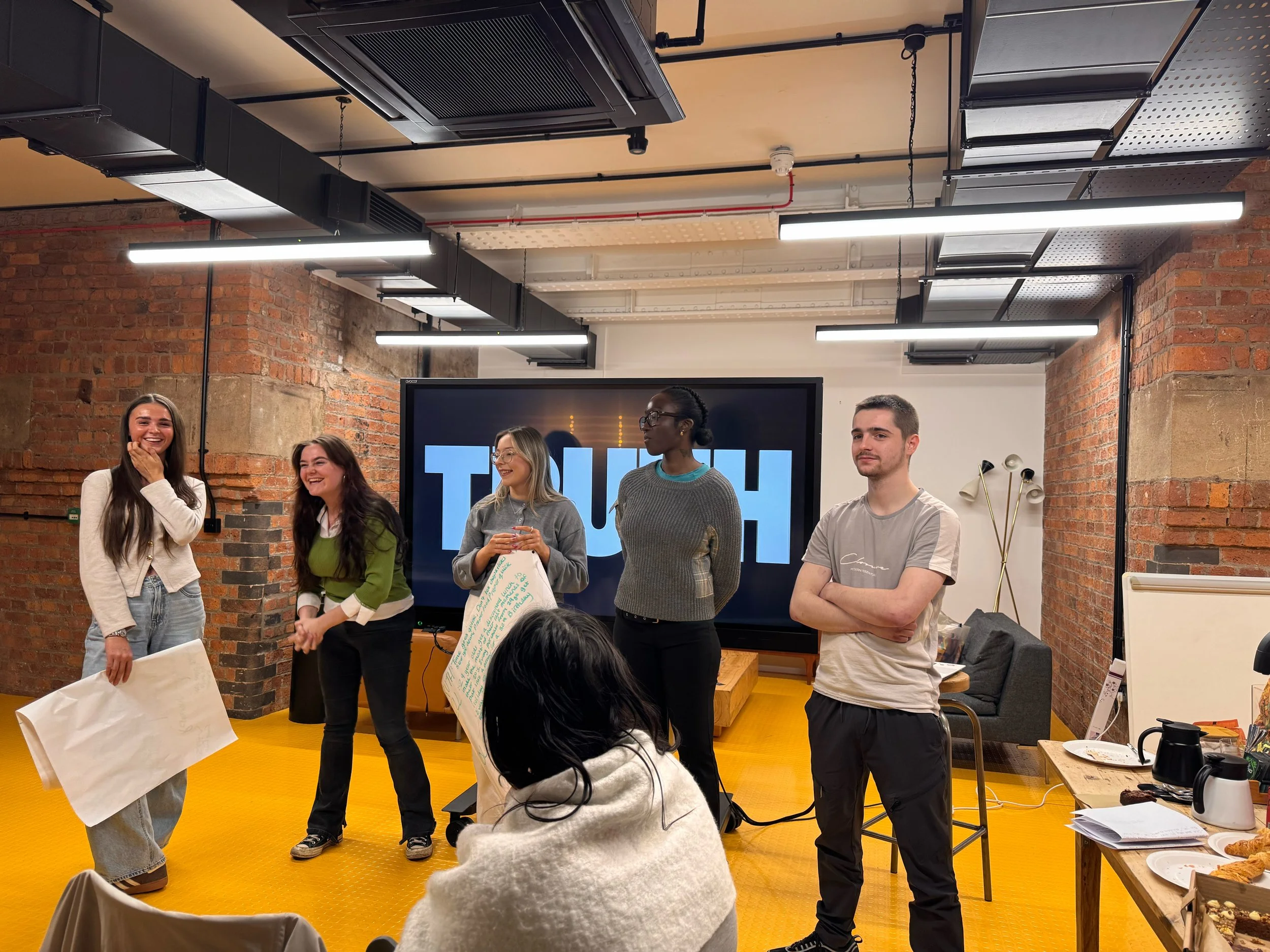 A group of six young adults standing in a room with exposed brick walls, black ceiling fixtures, and a large screen displaying 'TOUCH' in the background. They are engaging in a presentation or discussion, with one person holding a large poster with handwritten notes, and others smiling or looking attentive. There are tables with papers and food items, suggesting a casual meeting or workshop setting.