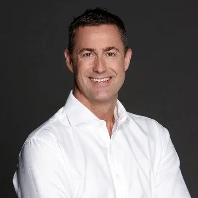 A man with short dark hair wearing a white dress shirt, smiling, against a dark gray background.