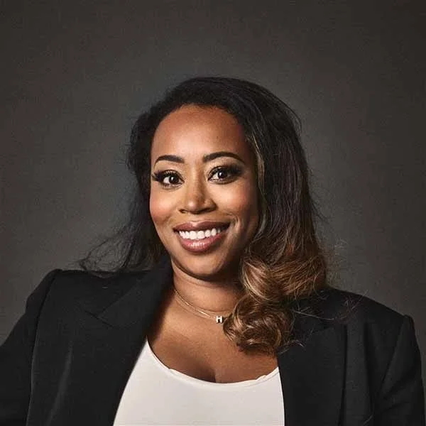 Portrait of a woman with wavy hair, wearing a black blazer and white top, smiling against a dark gray background.