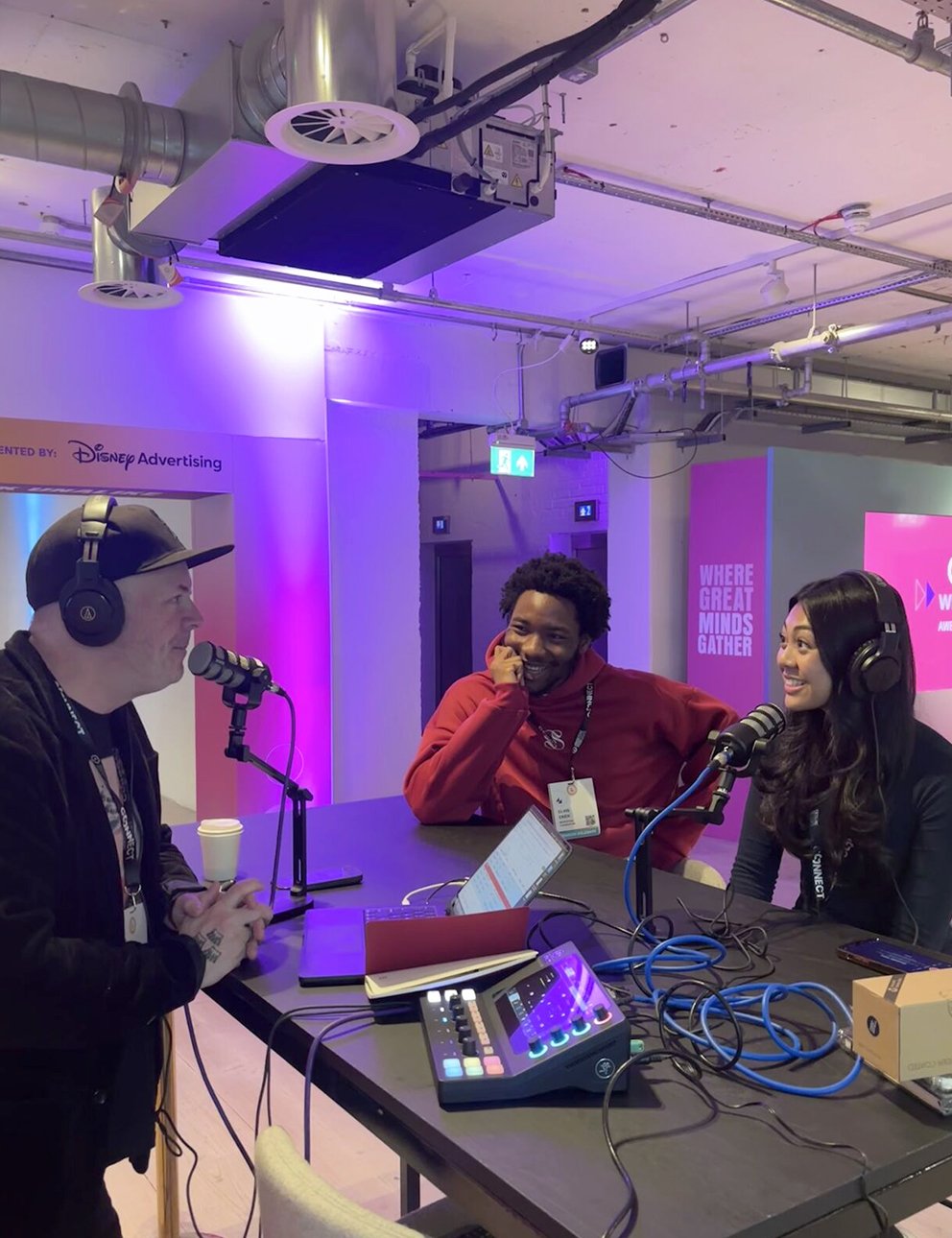 Three people sitting at a table with microphones and audio equipment, recording a podcast or radio show, in a brightly lit room with purple and pink lighting and industrial ceiling pipes.