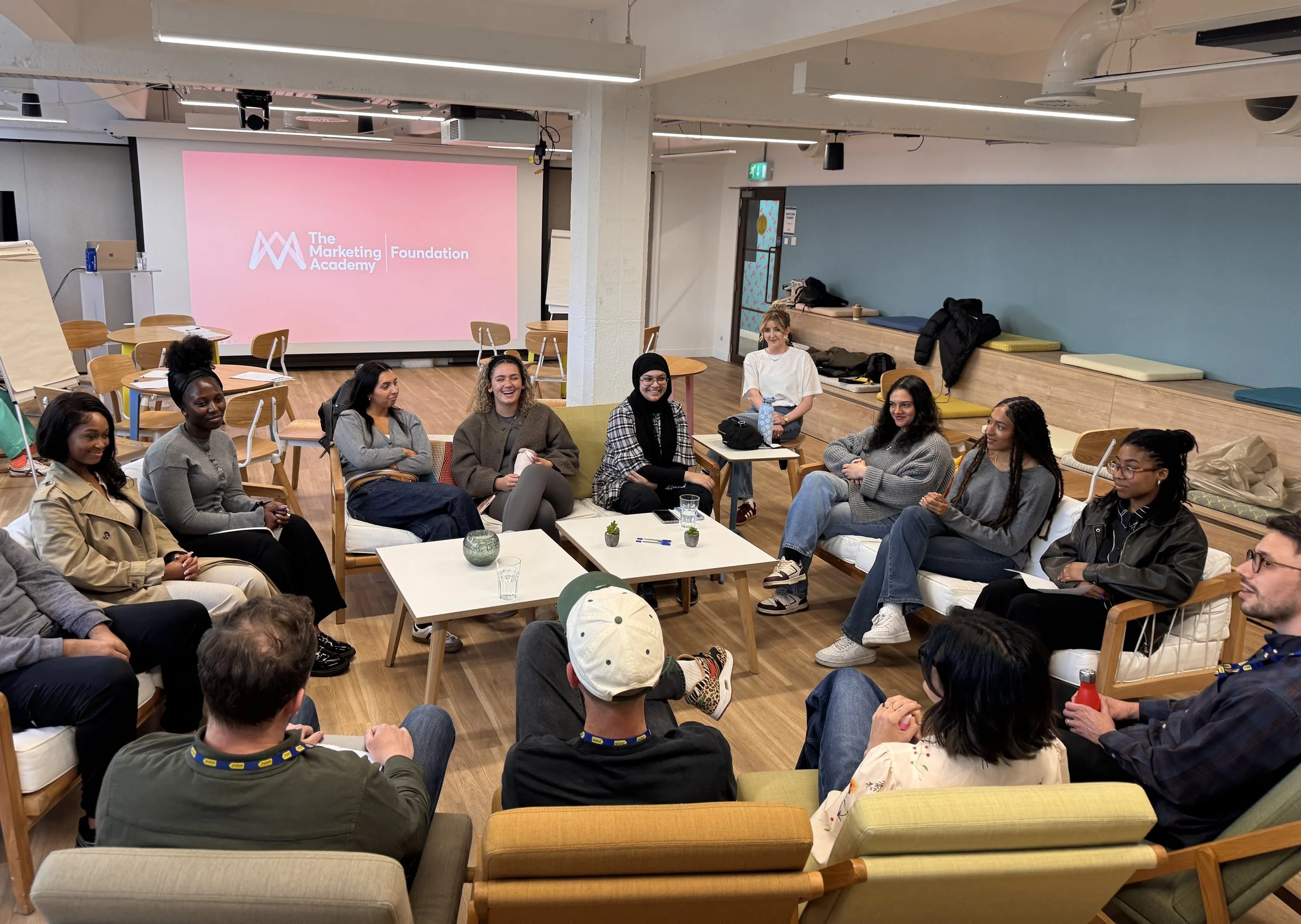 A group of diverse women and men attending a workshop or seminar in a modern, well-lit room. There are chairs and tables, with some people sitting on sofas and others standing or sitting on the floor. A large pink screen in the background displays the logo and text for 'The Marketing Academy Foundation.' The atmosphere appears friendly and engaging.