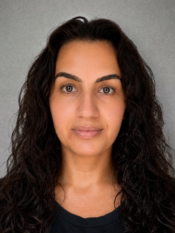 Close-up portrait of a woman with curly dark hair, brown eyes, and a neutral expression, wearing a black top against a grey background.