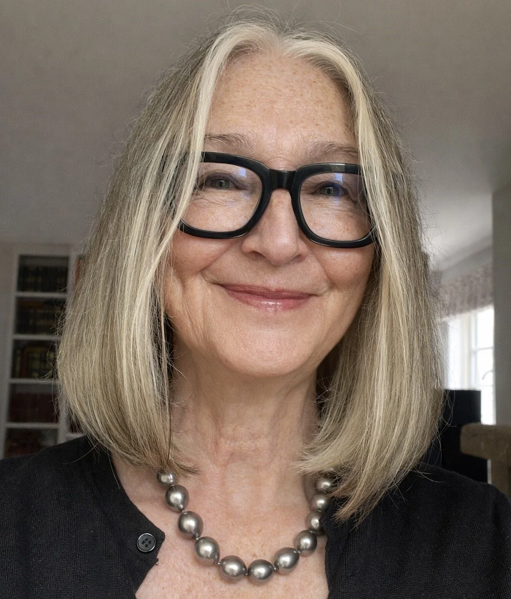 A smiling woman with shoulder-length blonde hair, black glasses, a pearl necklace, and a black top standing inside a room with a bookshelf and windows in the background.