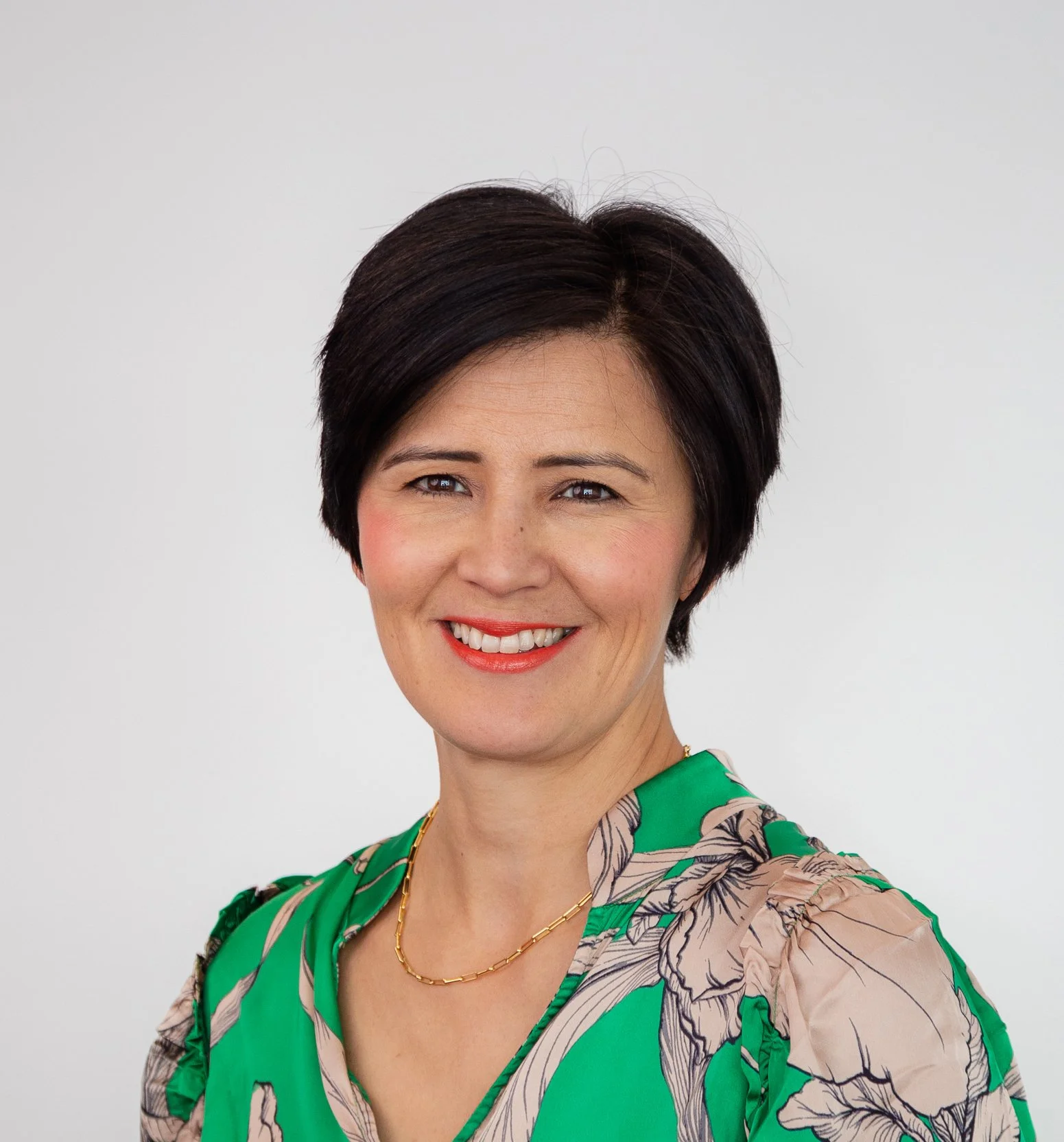 A woman with short dark hair, wearing a green dress with a floral pattern, gold necklace, and red lipstick, smiling against a plain white background.