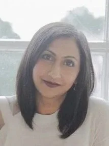 A woman with shoulder-length black hair wearing a white top, smiling softly, with a window in the background.