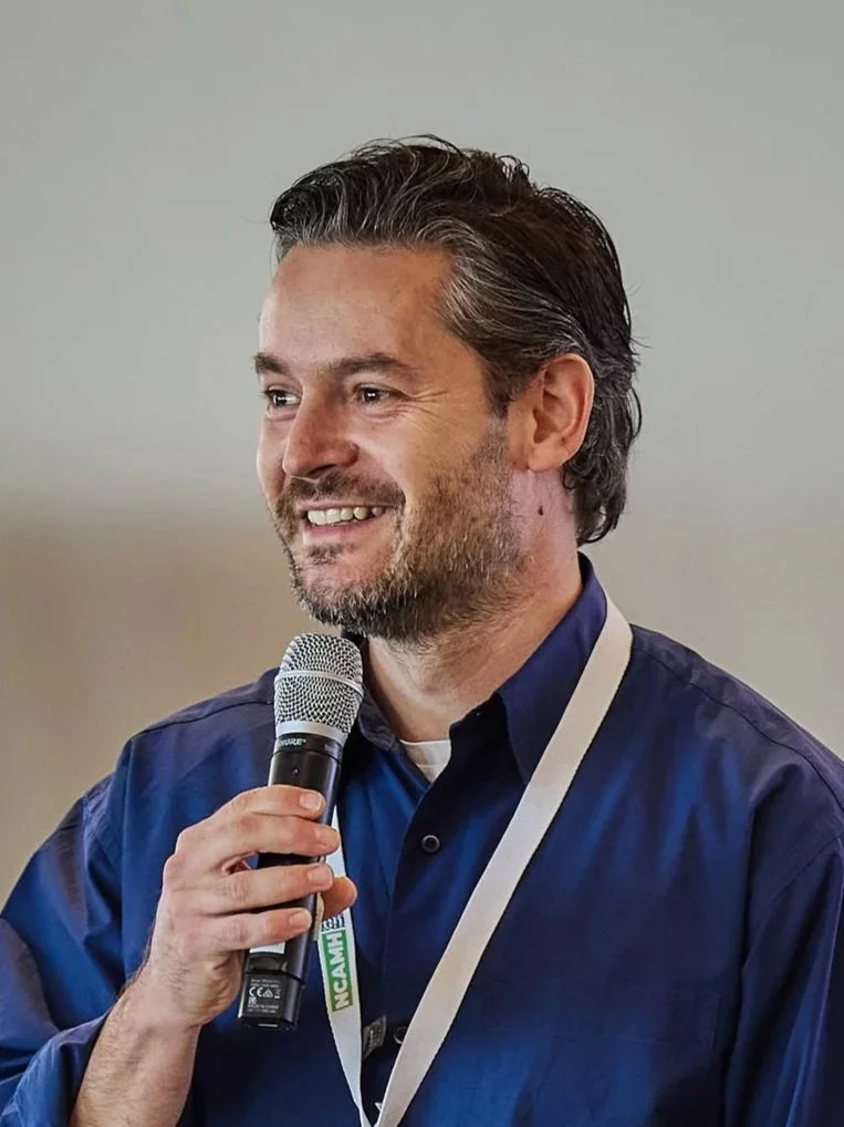 A man with dark hair and a beard, smiling, holding a microphone, wearing a navy blue shirt and a lanyard, speaking at an event.
