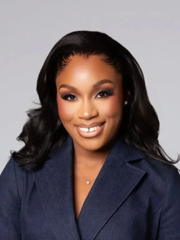 A smiling woman with shoulder-length black hair wearing a navy blazer and a delicate necklace, posing against a light gray background.