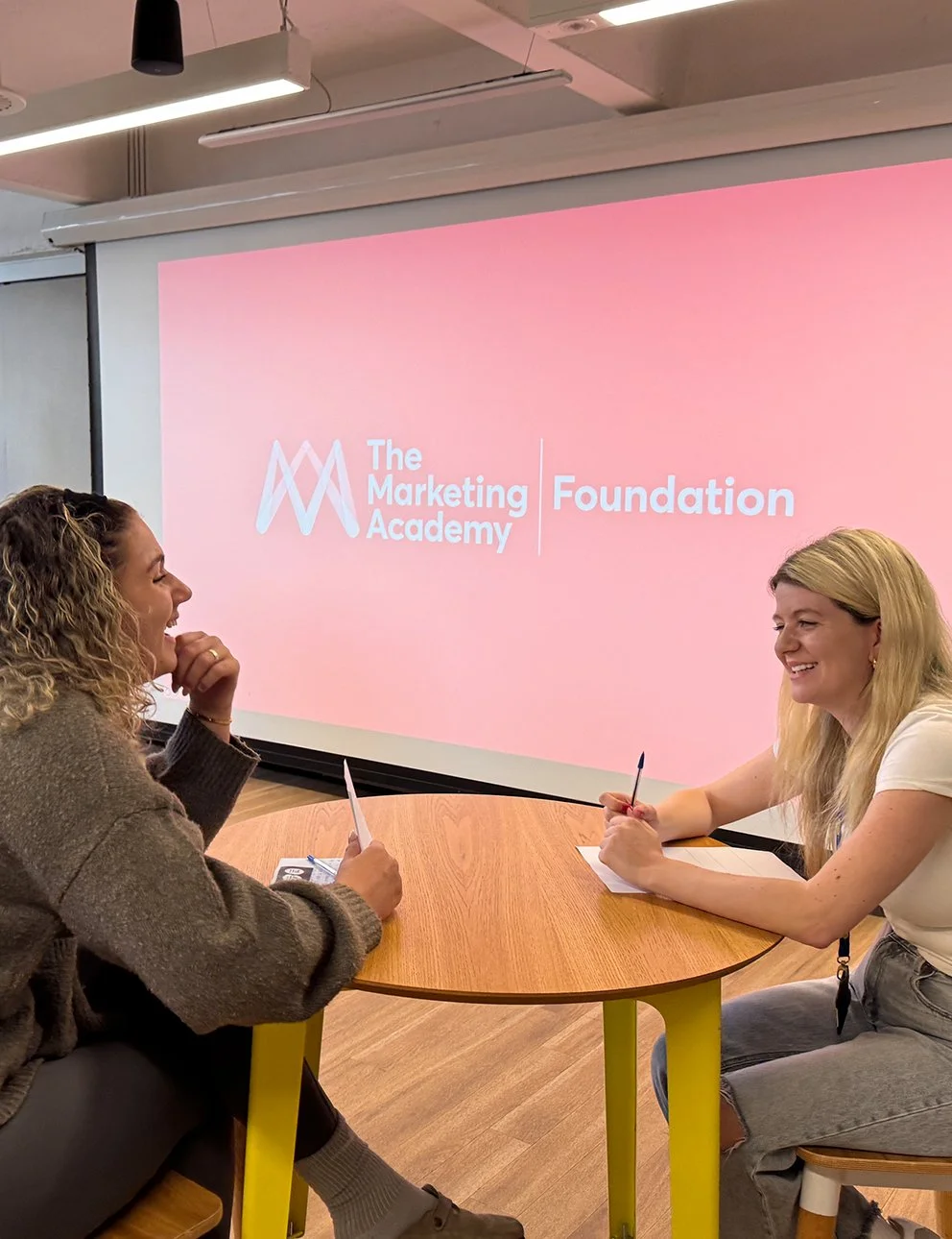 Two women sitting at a round table in a meeting room, engaged in conversation, with a large pink screen behind them displaying 'The Marketing Foundation' logo and text.