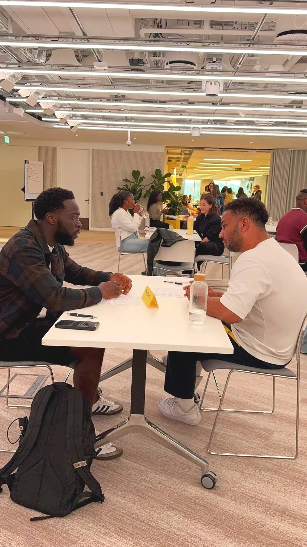 Two men sit at a table engaged in a conversation, with papers and a water bottle on the table. Other people are seated at tables in the background within a modern, well-lit room.