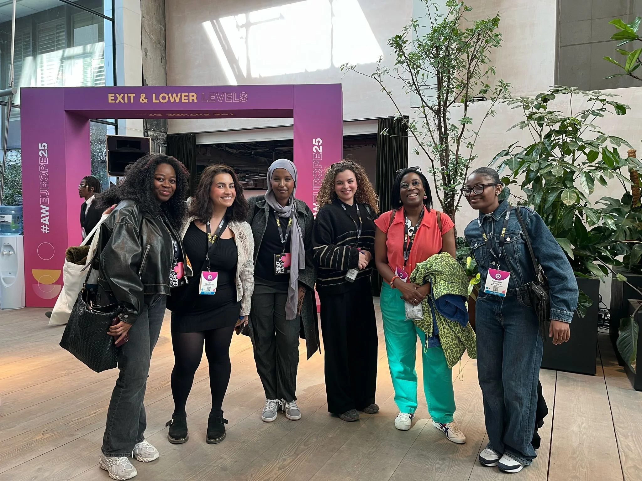 A group of six women standing together indoors in front of a black stage area with a purple banner above that reads 'EXIT & LOWER LEVELS'. They are smiling and wearing conference badges, with plants and large windows in the background.