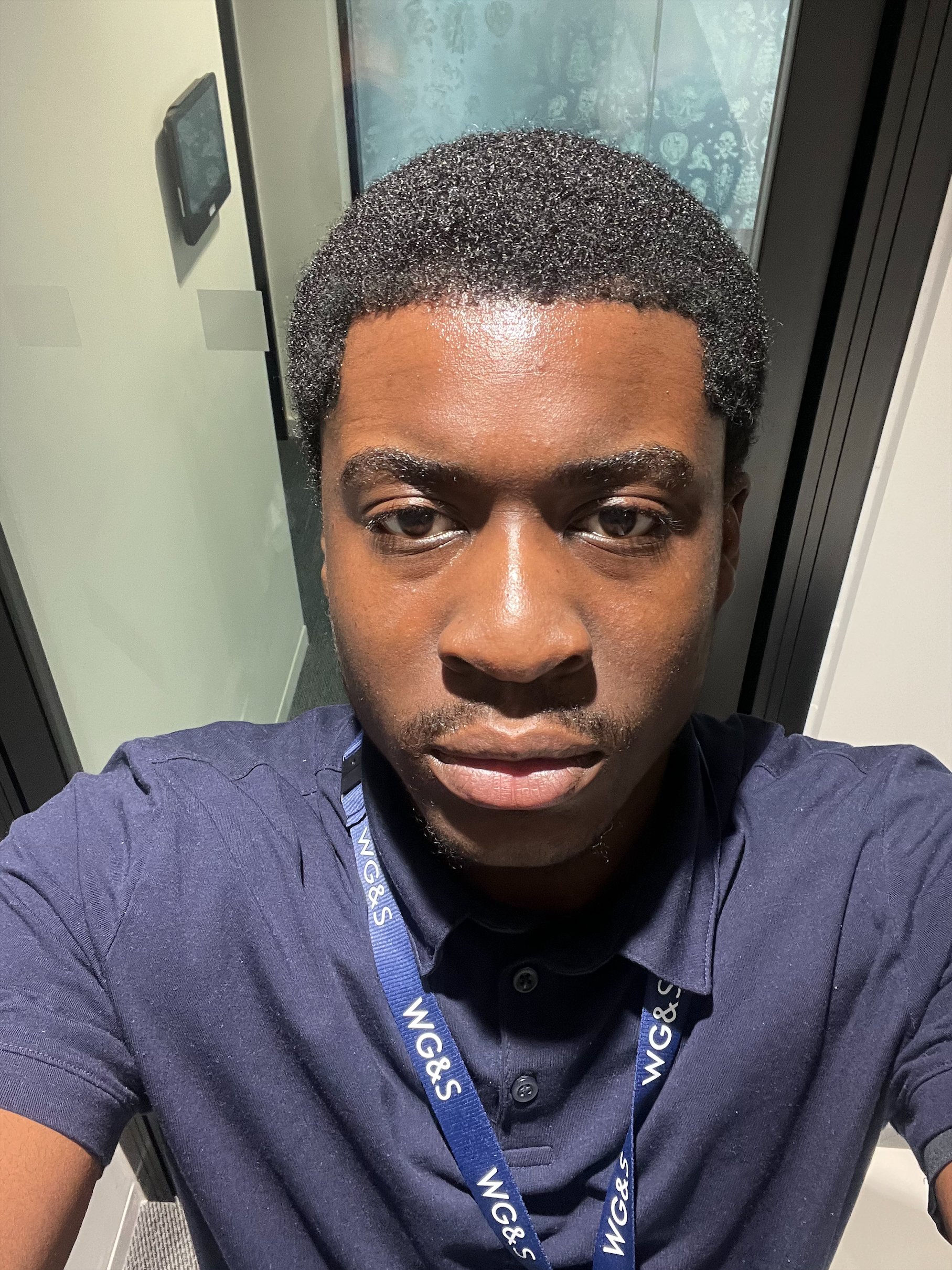 A man with short black curly hair, dark skin, and wearing a navy blue shirt takes a selfie indoors, sitting in front of a window with a reflection of plants and a door visible.