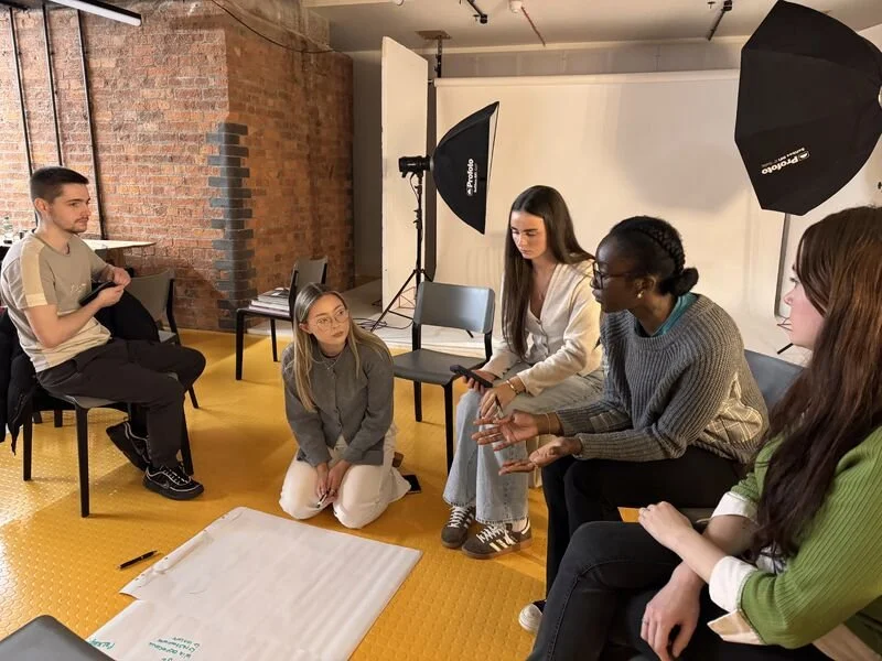 A group of five young women and one young man participating in a discussion or workshop in a room with brick walls and photography equipment. The women are seated, and the man is sitting on a chair, all with attentive expressions. There are lighting umbrellas and a whiteboard or paper on the floor with markers.