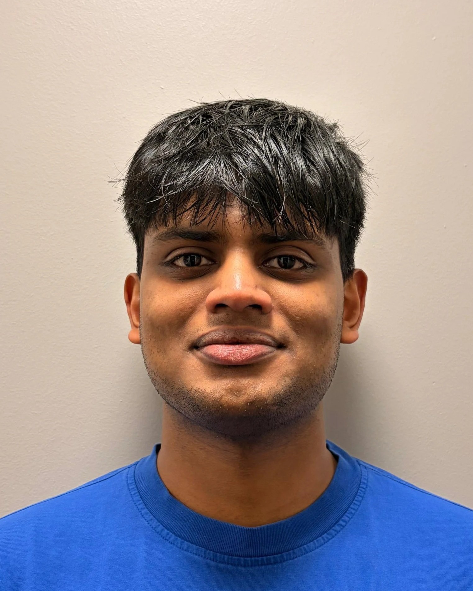 A young man with short dark hair, dark eyes, and tan skin wearing a blue shirt, standing against a plain beige wall.