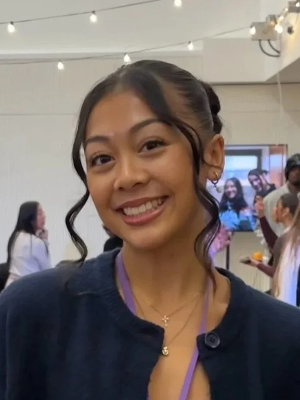 Smiling young woman with dark hair in an updo with loose curls, wearing a black top, gold jewelry, and a purple lanyard, at an indoor event with other people and string lights.