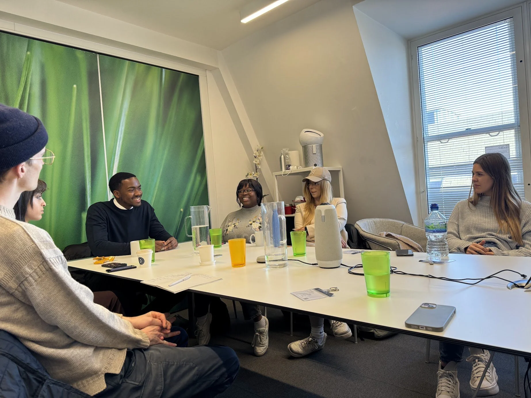 A diverse group of six people sitting around a white conference table in a modern office, engaged in discussion. The table holds colorful cups, a smartphone, papers, and a projector. The room has a large green wall or mural, with windows and blinds on the right side, and a shelf with a humidifier and other items in the background.