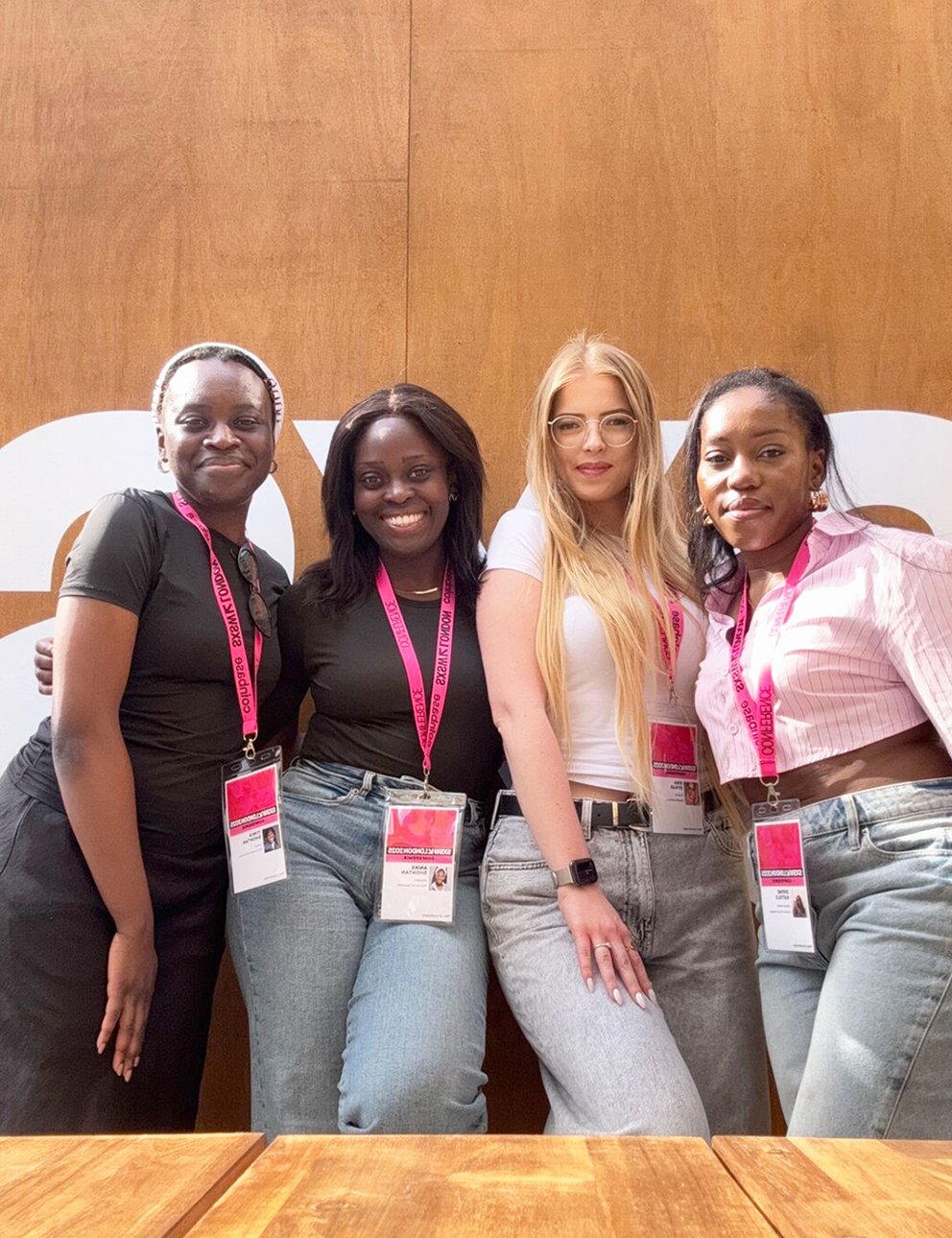 Four women standing together at a conference, wearing name badges and pink lanyards, in front of a wooden wall with large white letters behind them.