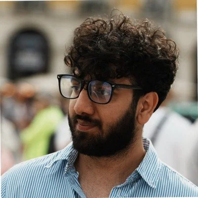 A man with curly hair, glasses, and a beard wearing a light blue striped shirt, standing outdoors.