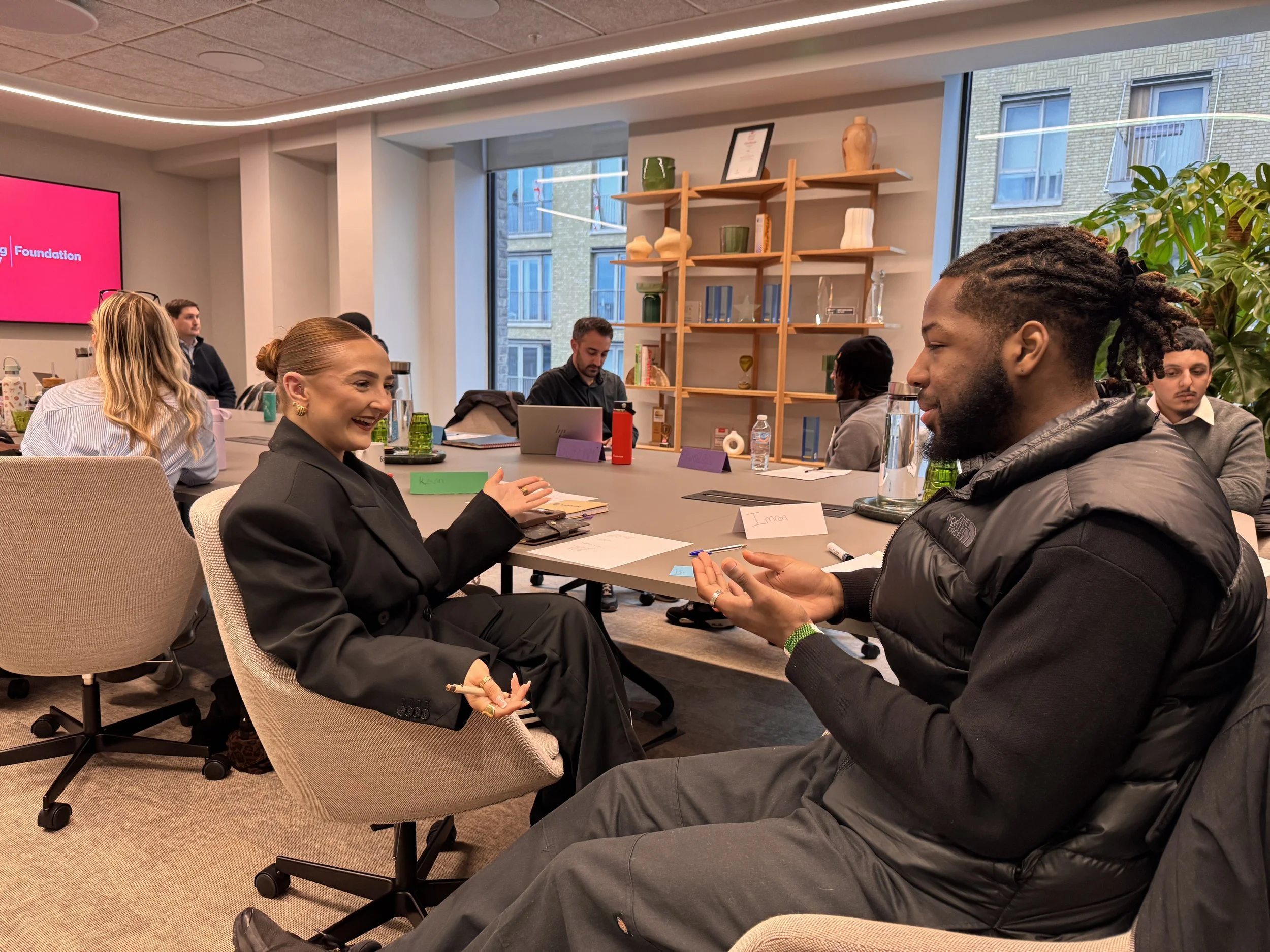 People sitting around a conference table having a discussion, some are smiling and others are listening attentively, in a well-lit room with large windows and decorative items on shelves.