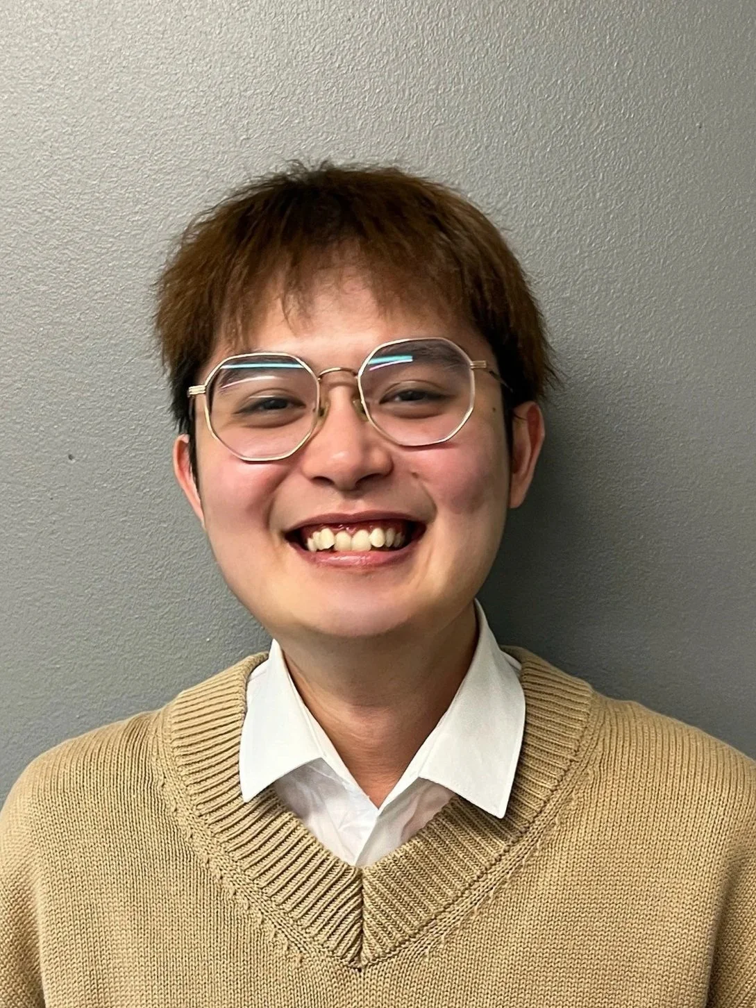 A young person smiling against a plain wall with glasses, short brown hair, a white collared shirt, and a beige sweater.