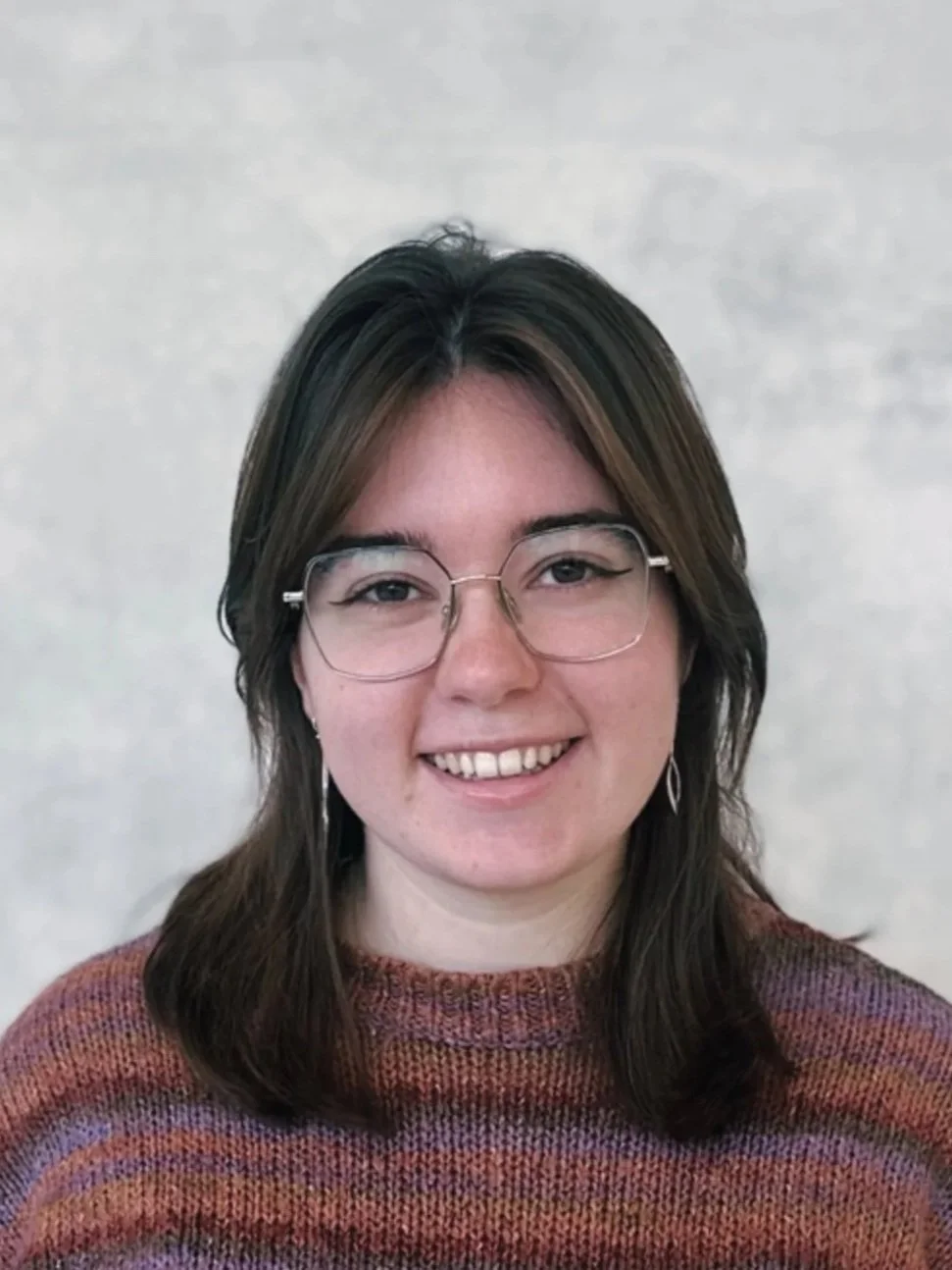 A woman with shoulder-length brown hair, glasses, and earrings, smiling at the camera, wearing a multi-colored sweater, against a plain light background.