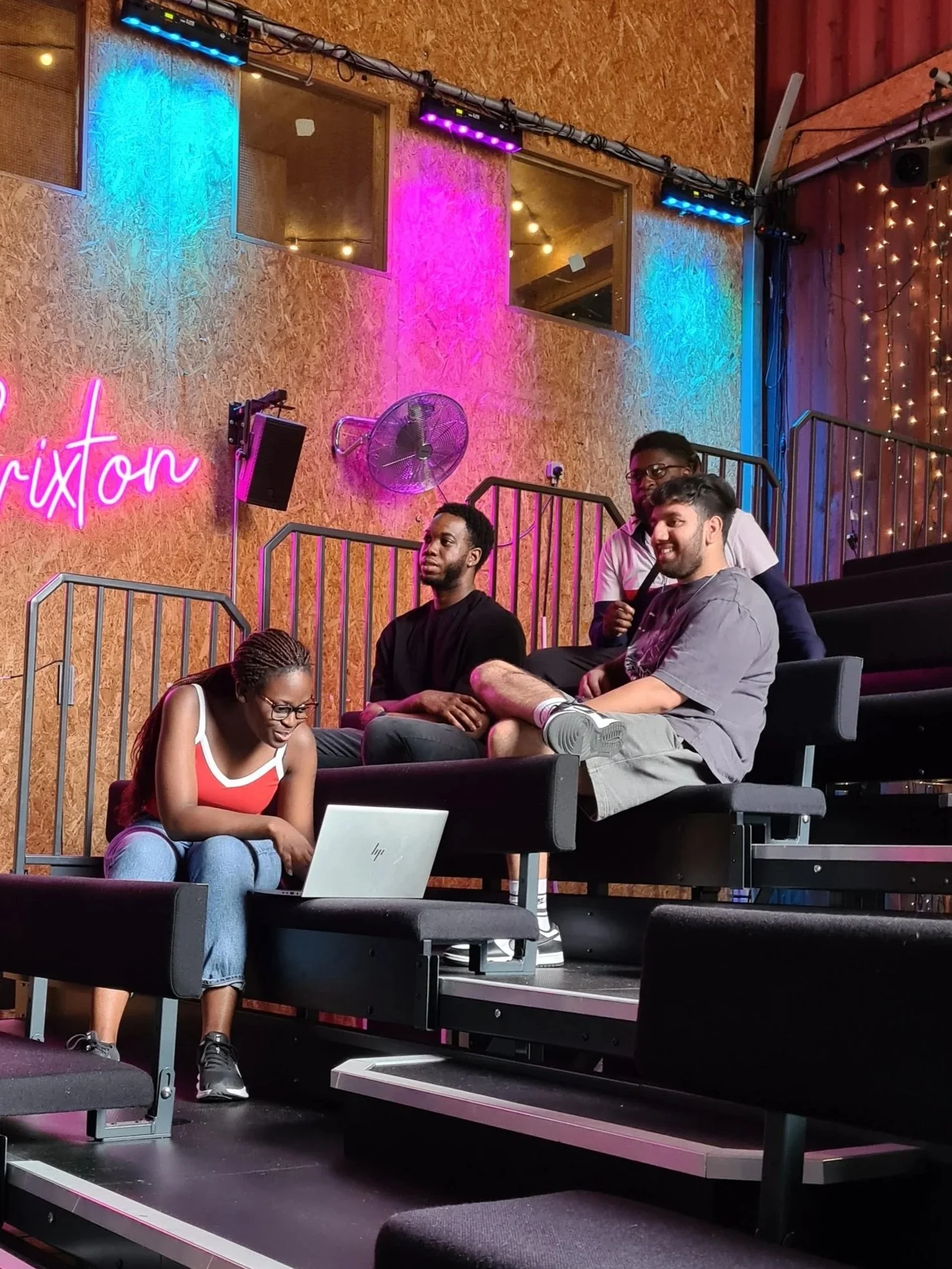 People sitting on tiered seats in a studio or auditorium with colorful lighting, a neon sign, and a computer in front of the woman in the foreground.